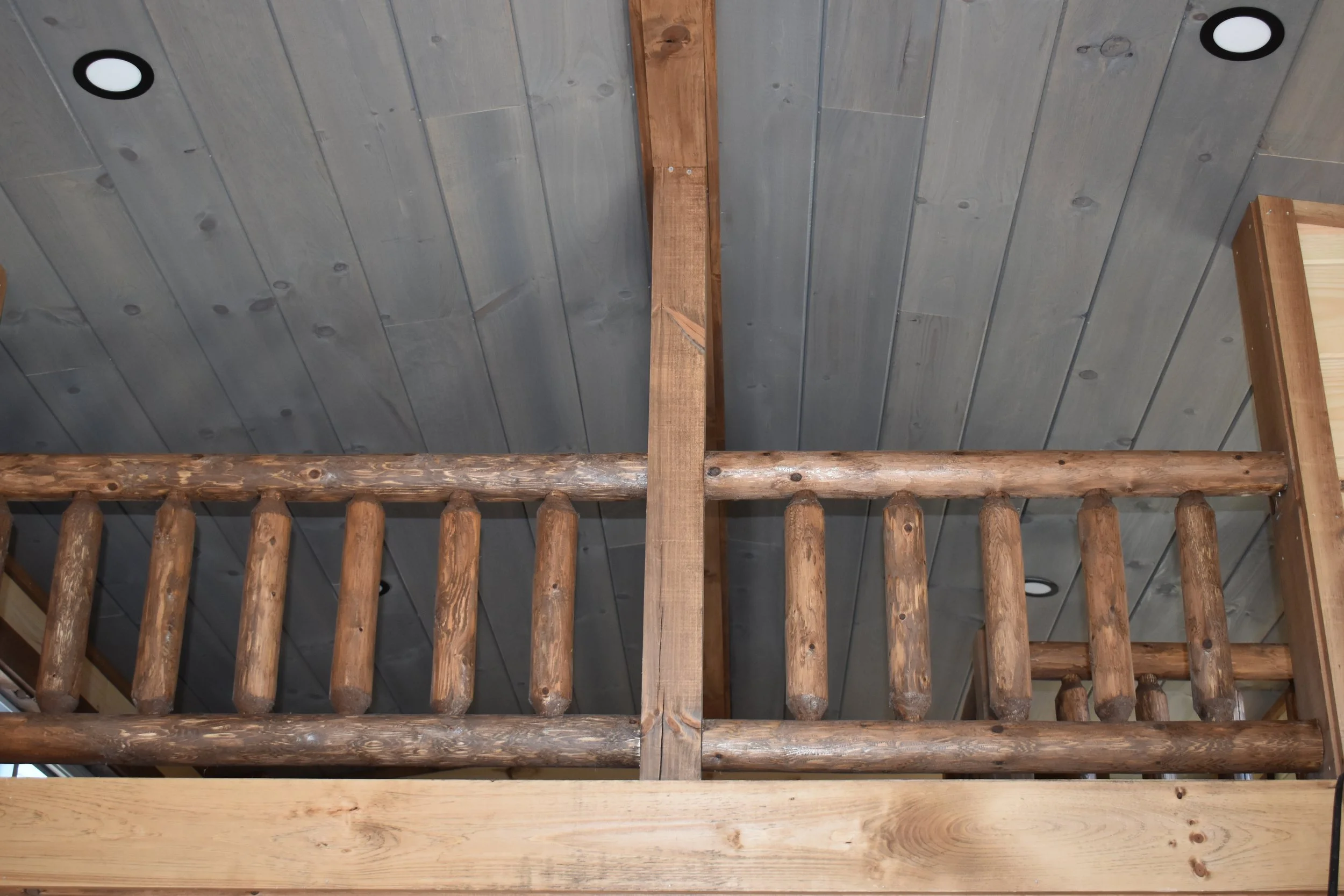 Interior view of a rustic wooden mezzanine or balcony with log railings and a wood-paneled ceiling with recessed lights.