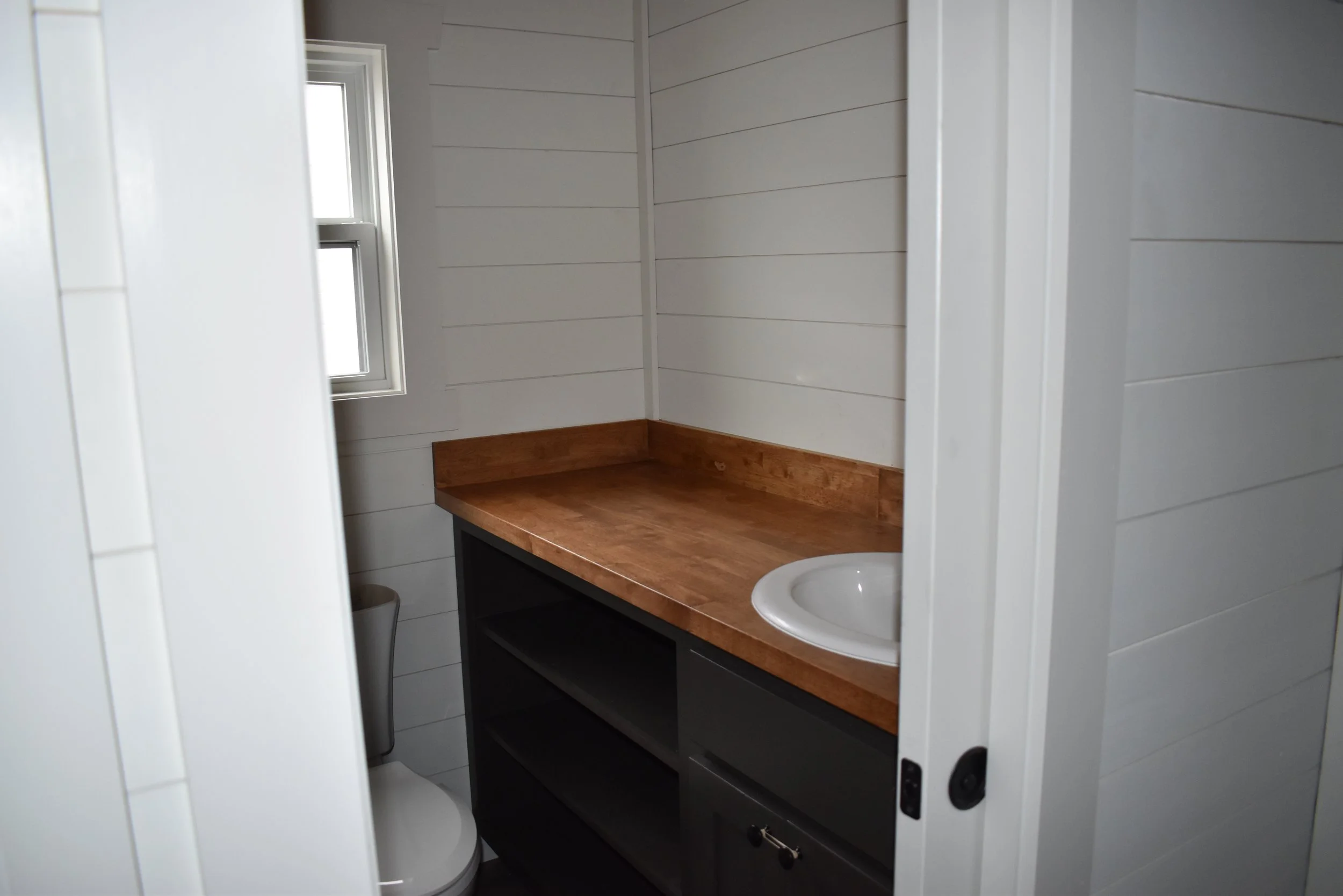 Small bathroom with a window, white shiplap walls, a wooden countertop with a white sink, black shelving unit, and a toilet partially visible.