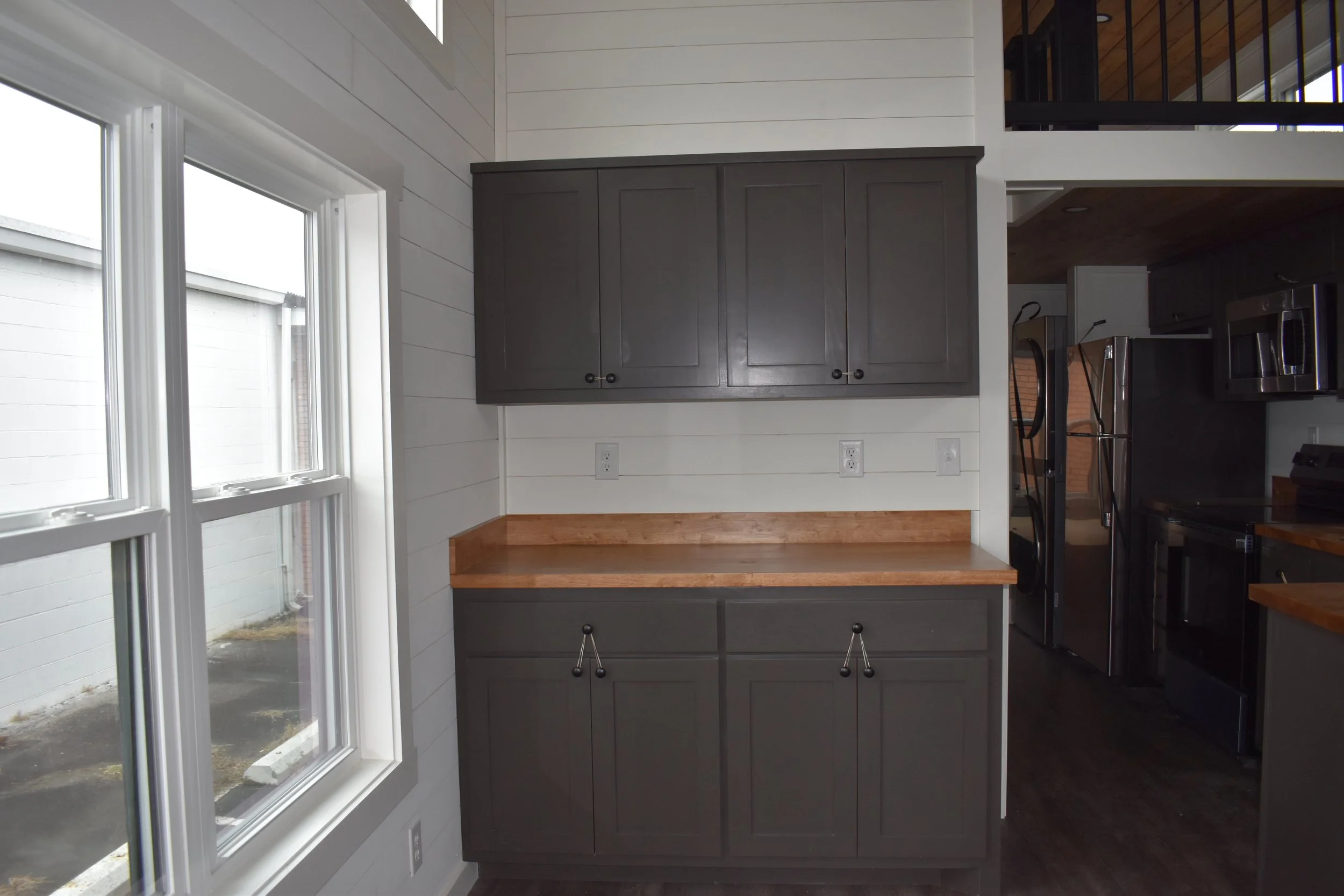 Kitchen corner with dark gray cabinets, wooden countertop, white paneled walls, window with double glass panes, and a view into other kitchen appliances.