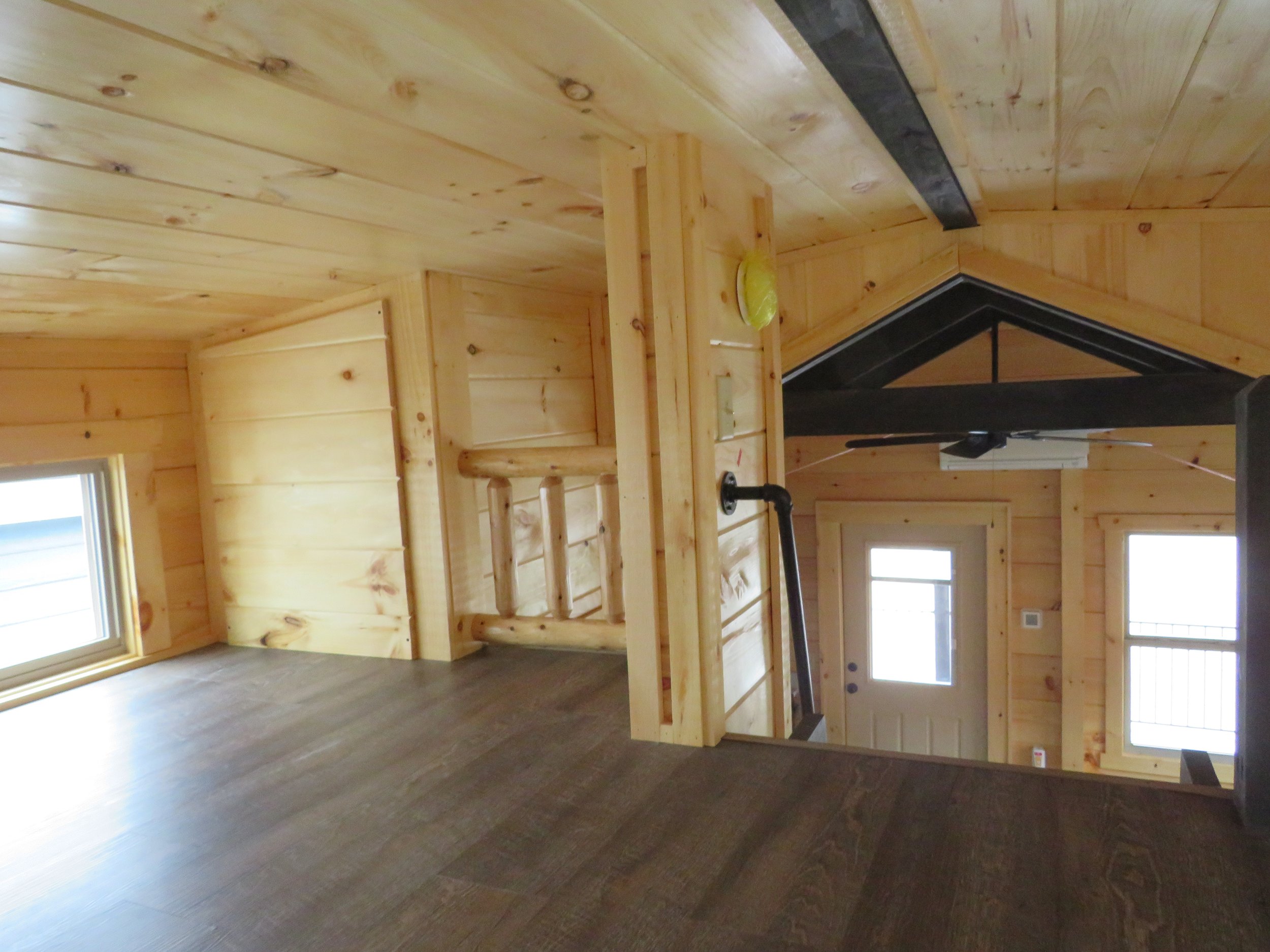 View of the interior of a house with wooden walls, ceiling, and staircase, showing a landing and staircase leading down, with windows and a door visible.
