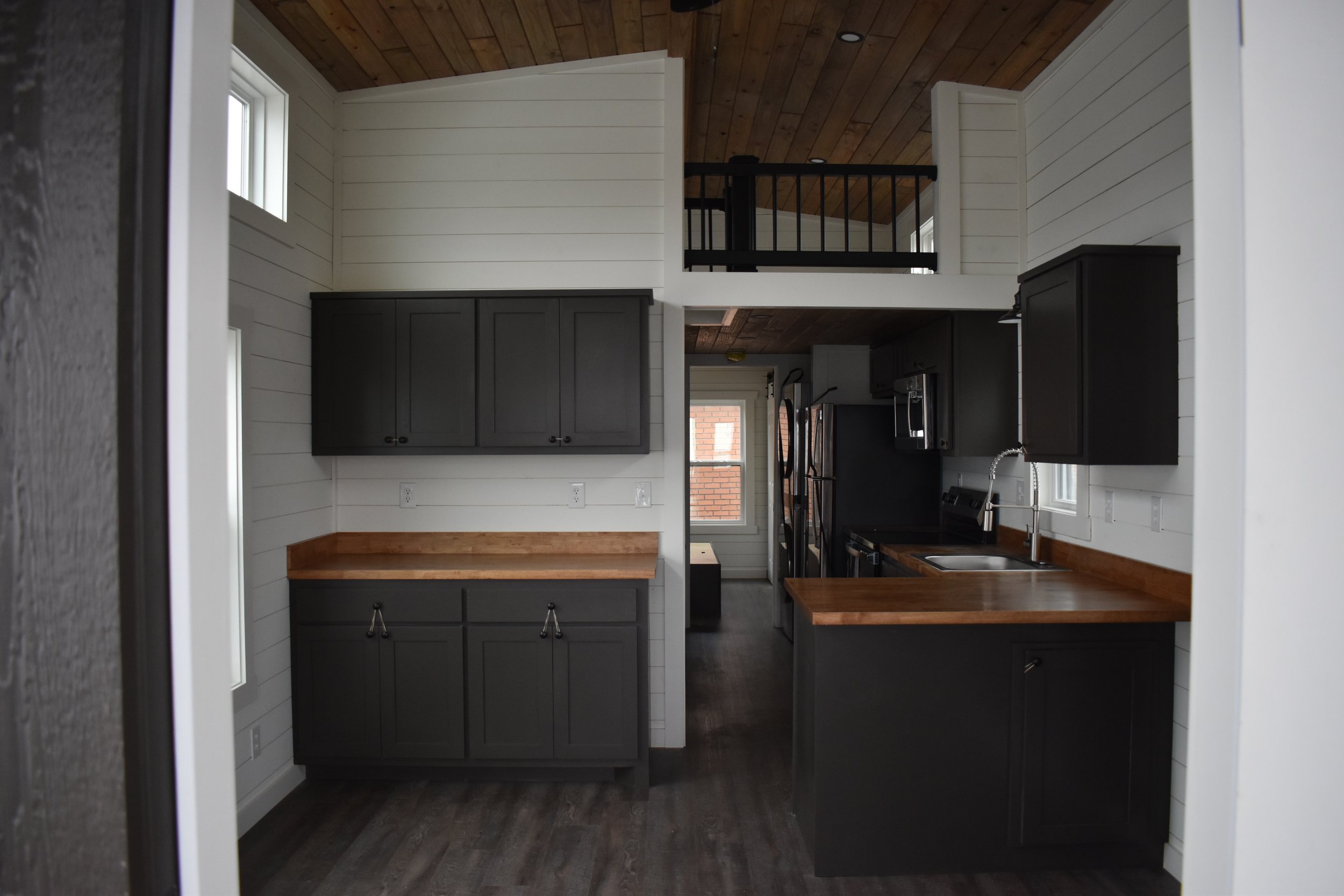 Modern kitchen with black cabinets, wooden countertops, white shiplap walls, and a vaulted ceiling with wood paneling, overlooking a living space with a loft area.