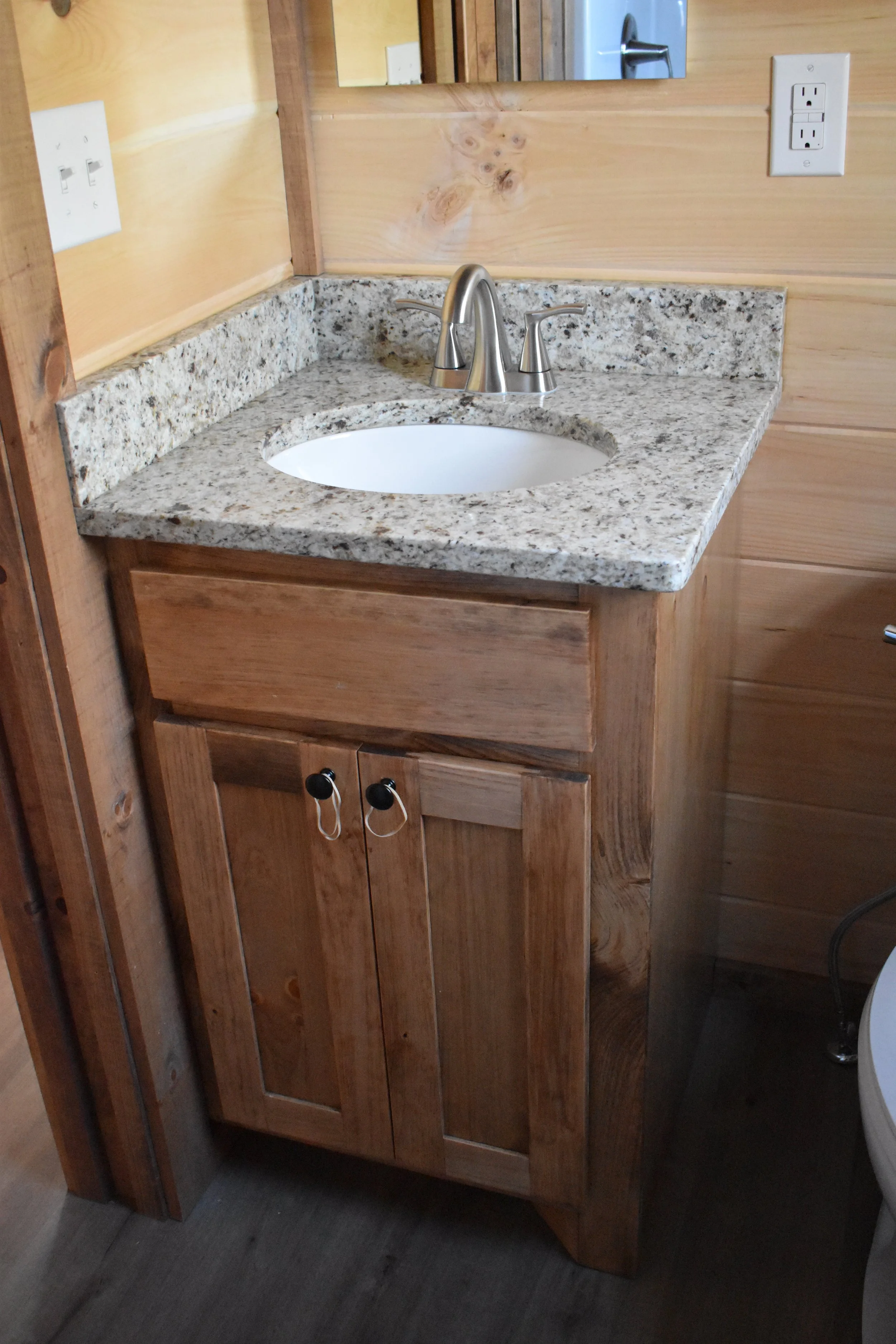 A bathroom vanity with a granite countertop, a white oval sink, and a brushed nickel faucet, against wooden paneled walls and a mirror above.