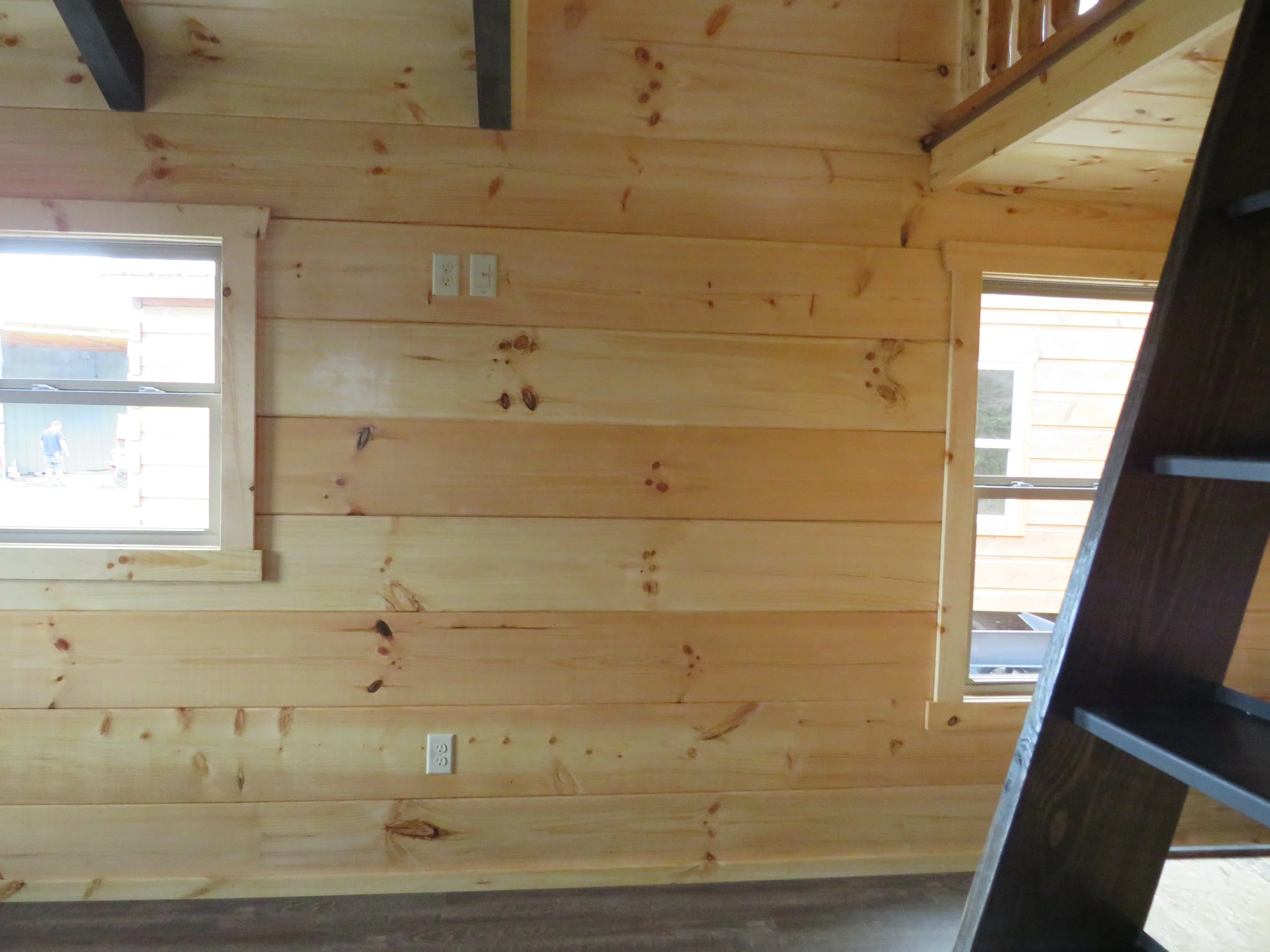 Wooden interior wall with two windows, electrical outlets, and a dark wooden staircase or railing in the foreground.