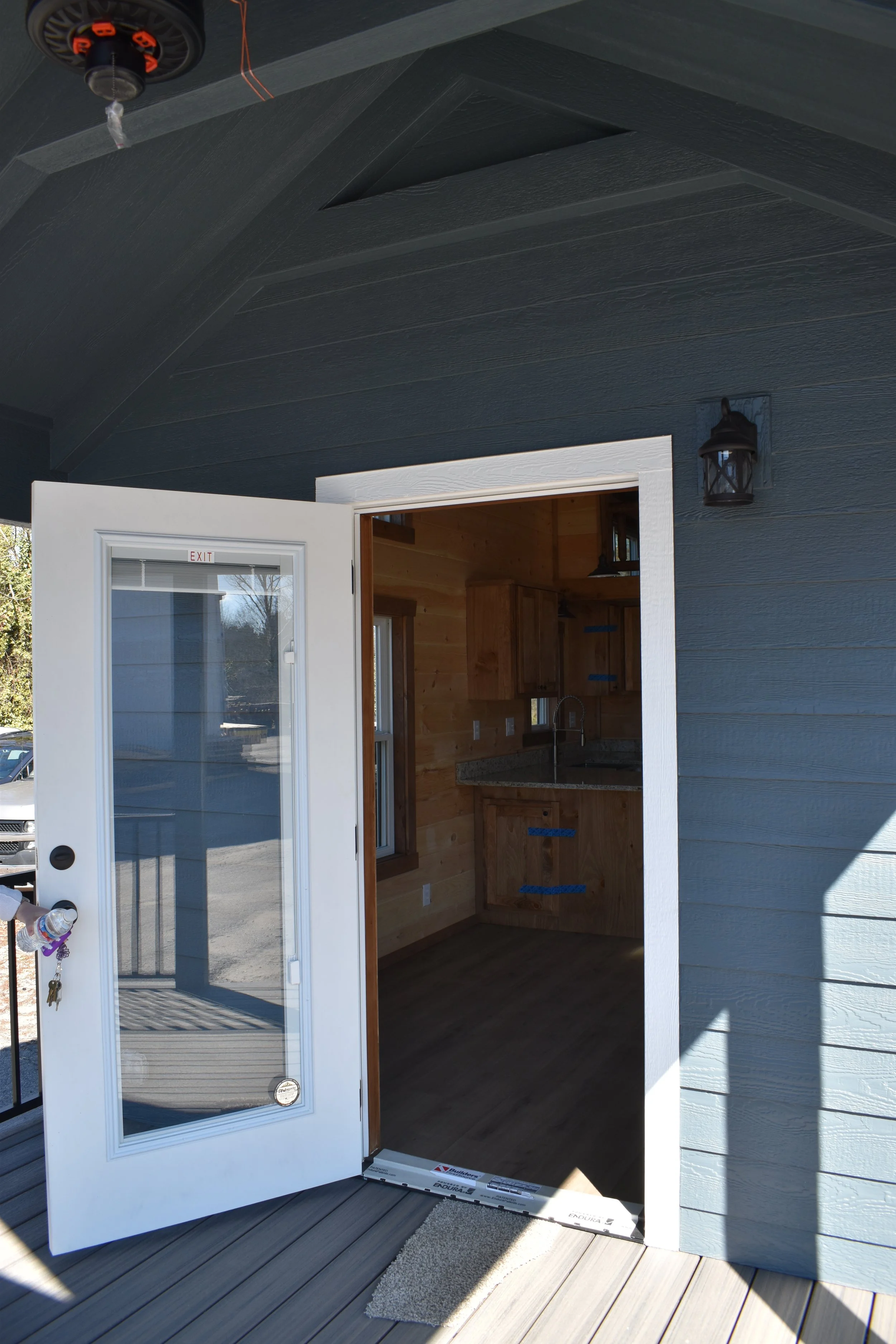 Exterior view of a partially open white door leading into a wooden interior room with a kitchen, visible cabinets, window, and countertop. The building has blue exterior walls, a dark ceiling, a wall-mounted lantern, and a small outdoor mat.