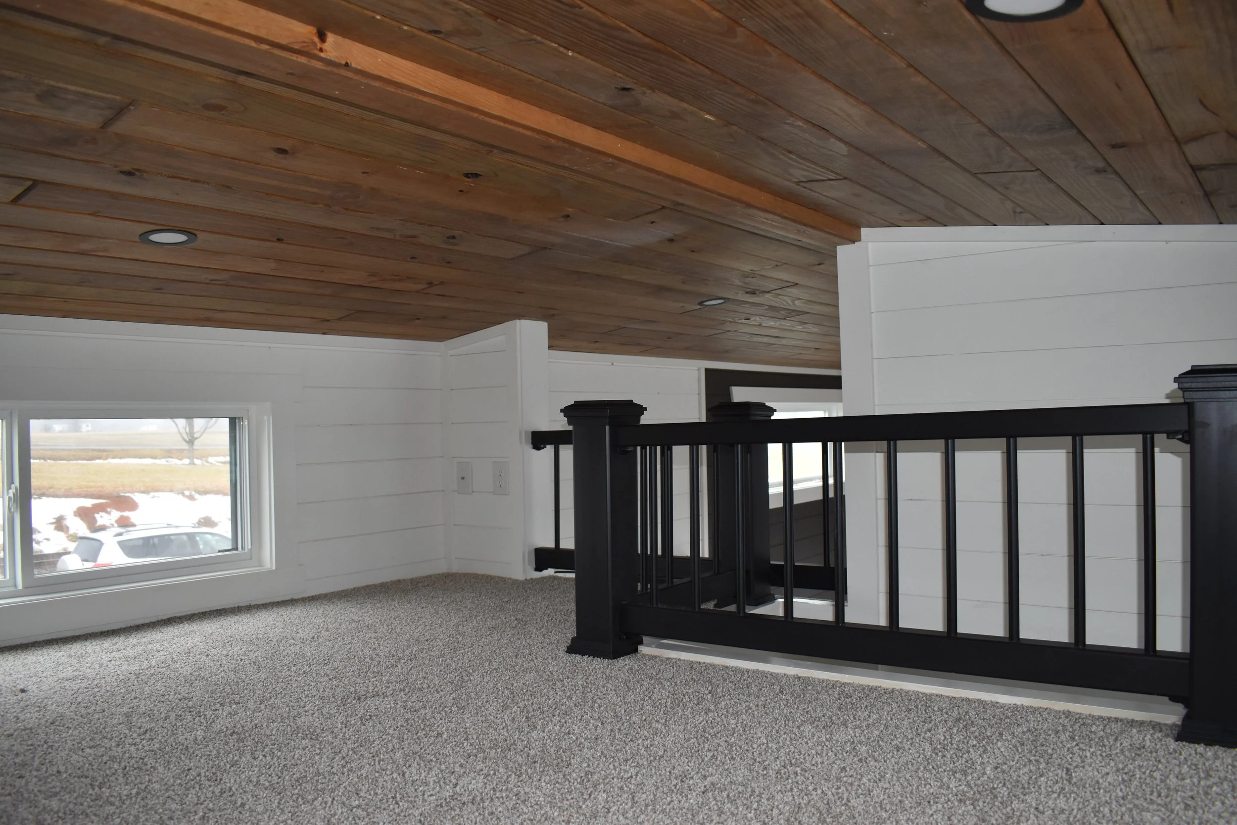 Interior view of a loft or upper room with cork ceiling, white shiplap walls, carpeted floor, a black railing, and a window showing an outdoor scene of snow and cars.