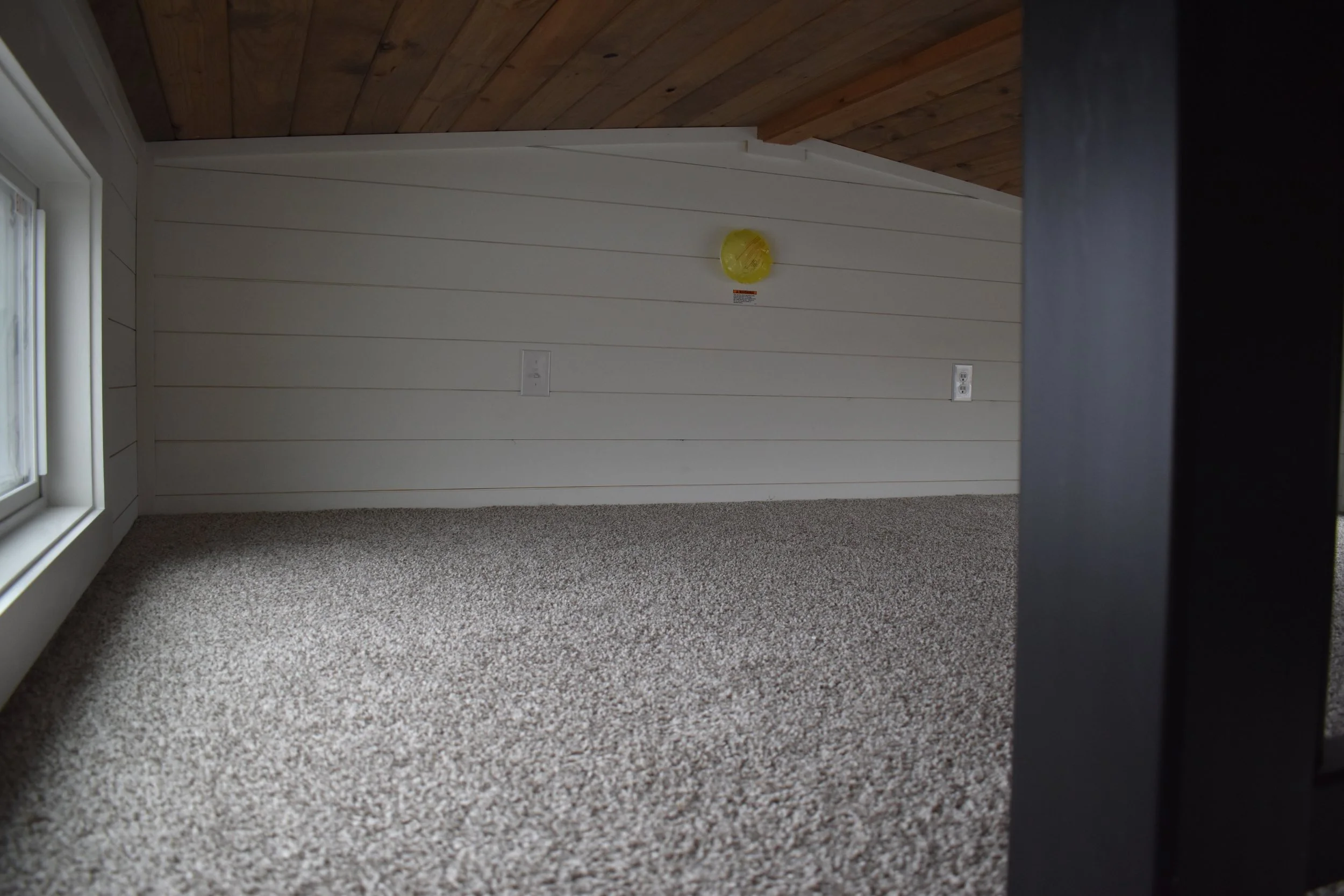 Empty attic space with beige carpet, white wall paneling, a small yellow object on the wall, two electrical outlets, a window on the left, and a wooden ceiling.