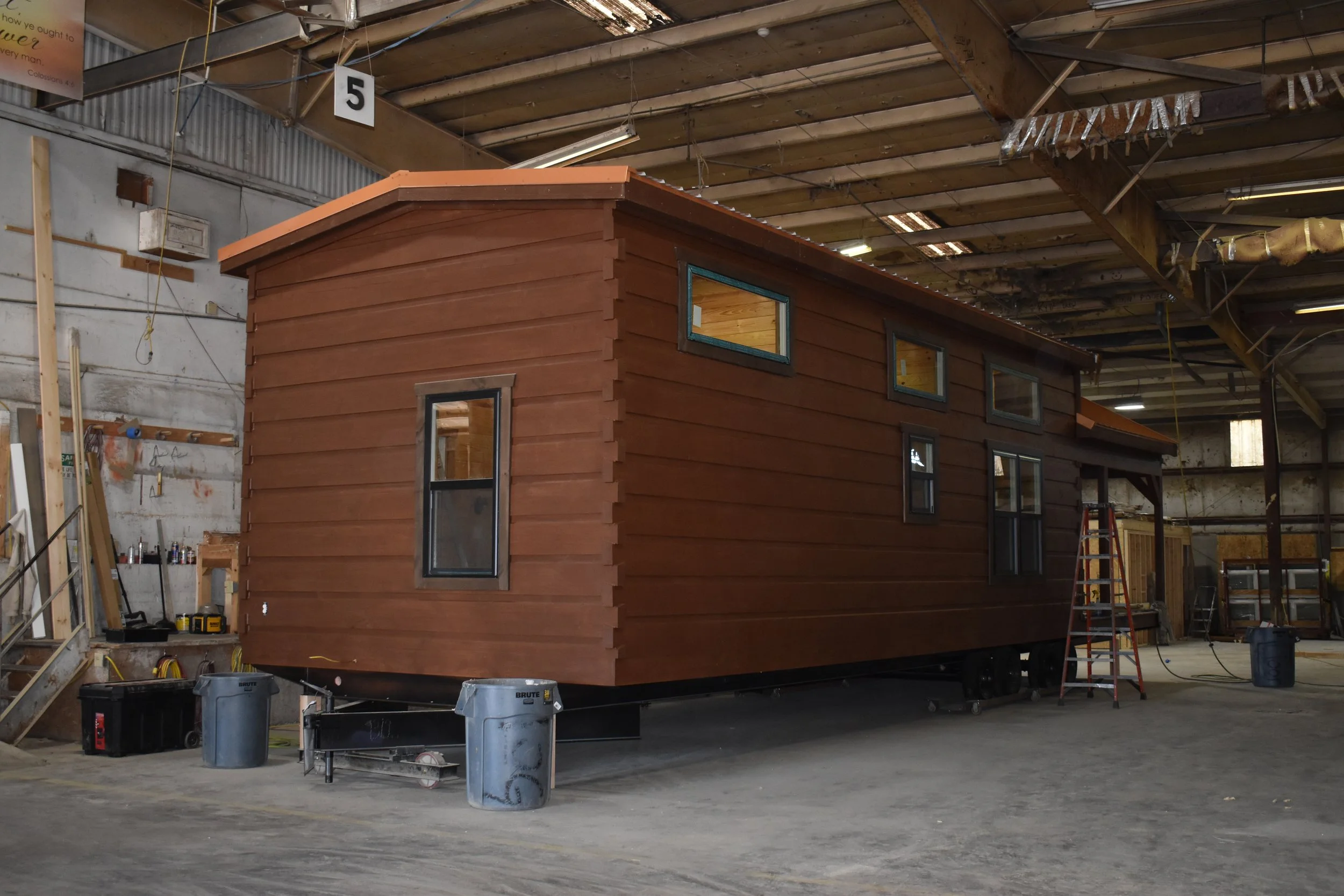 A small wooden house under construction inside a warehouse, with tools, bins, and construction materials around.