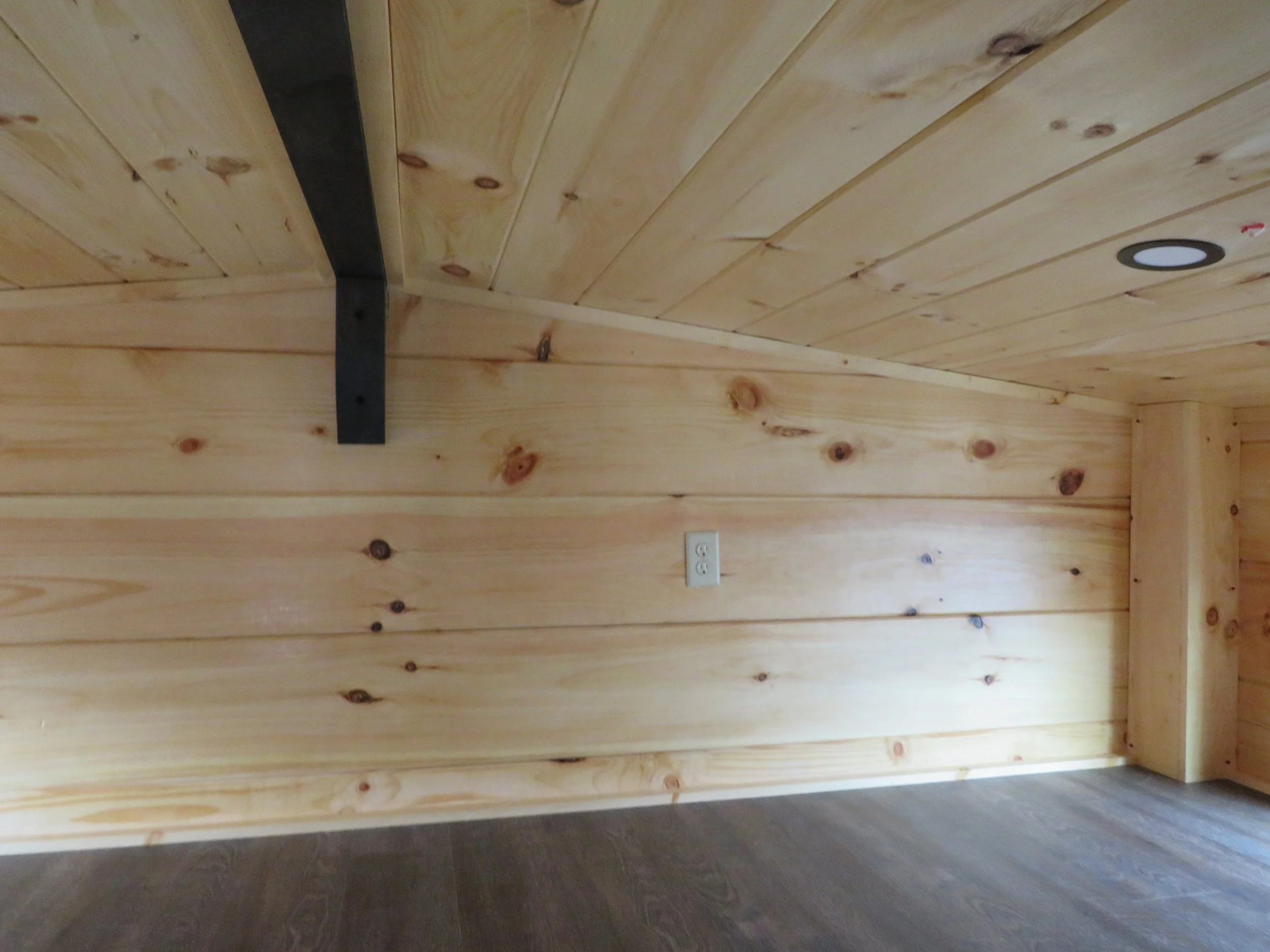 Wood-paneled wall with a black metal support bracket, a white electrical outlet, and a recessed ceiling light.