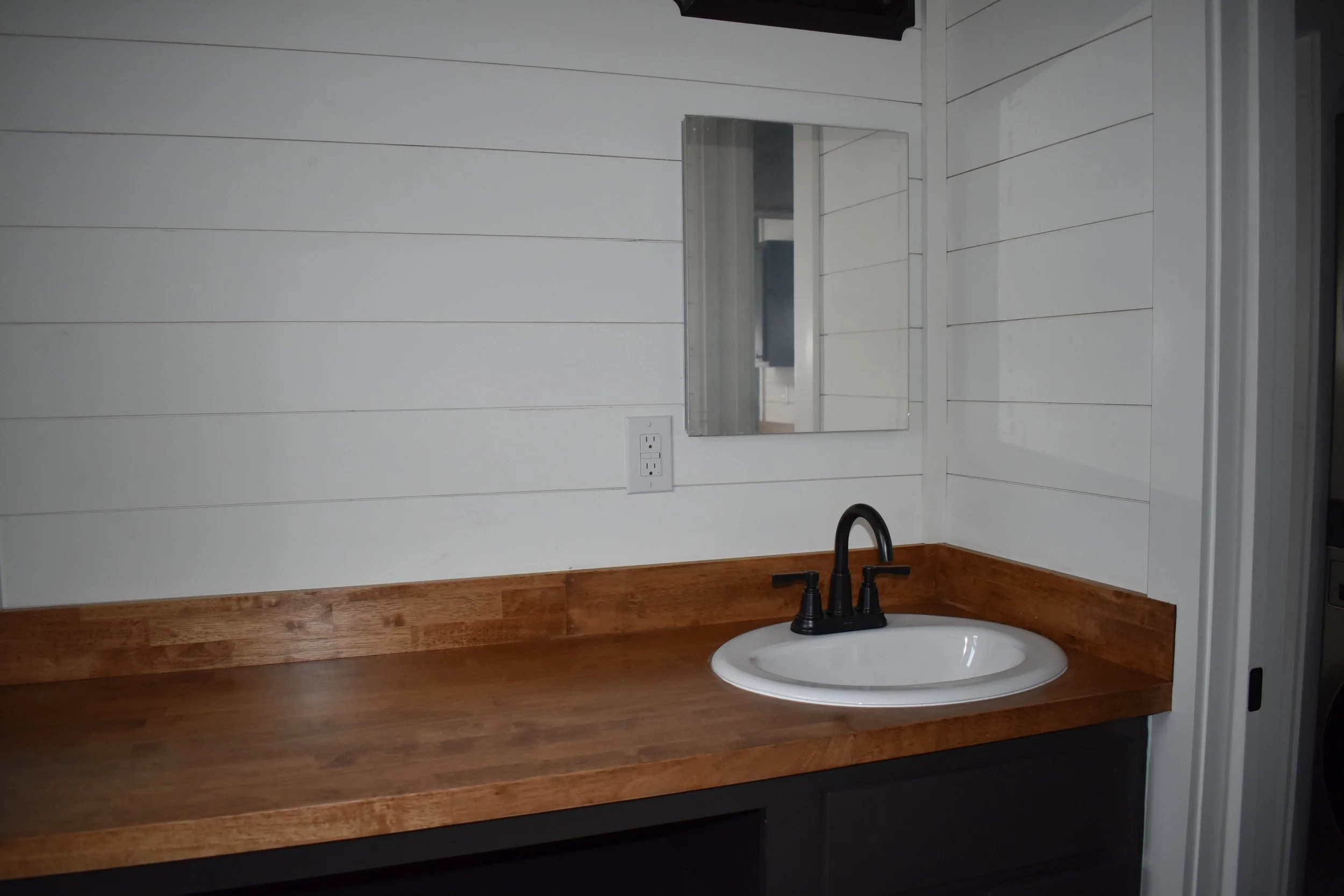 Bathroom vanity with a wooden countertop, a white oval sink, a black faucet, a mirror on a white paneled wall, and electrical outlets.