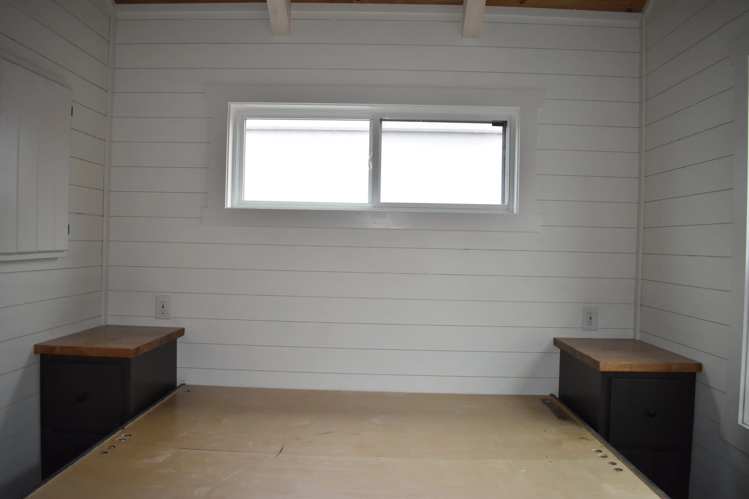 Empty room with white shiplap walls, a horizontal window, and two built-in black nightstands with wooden tops.