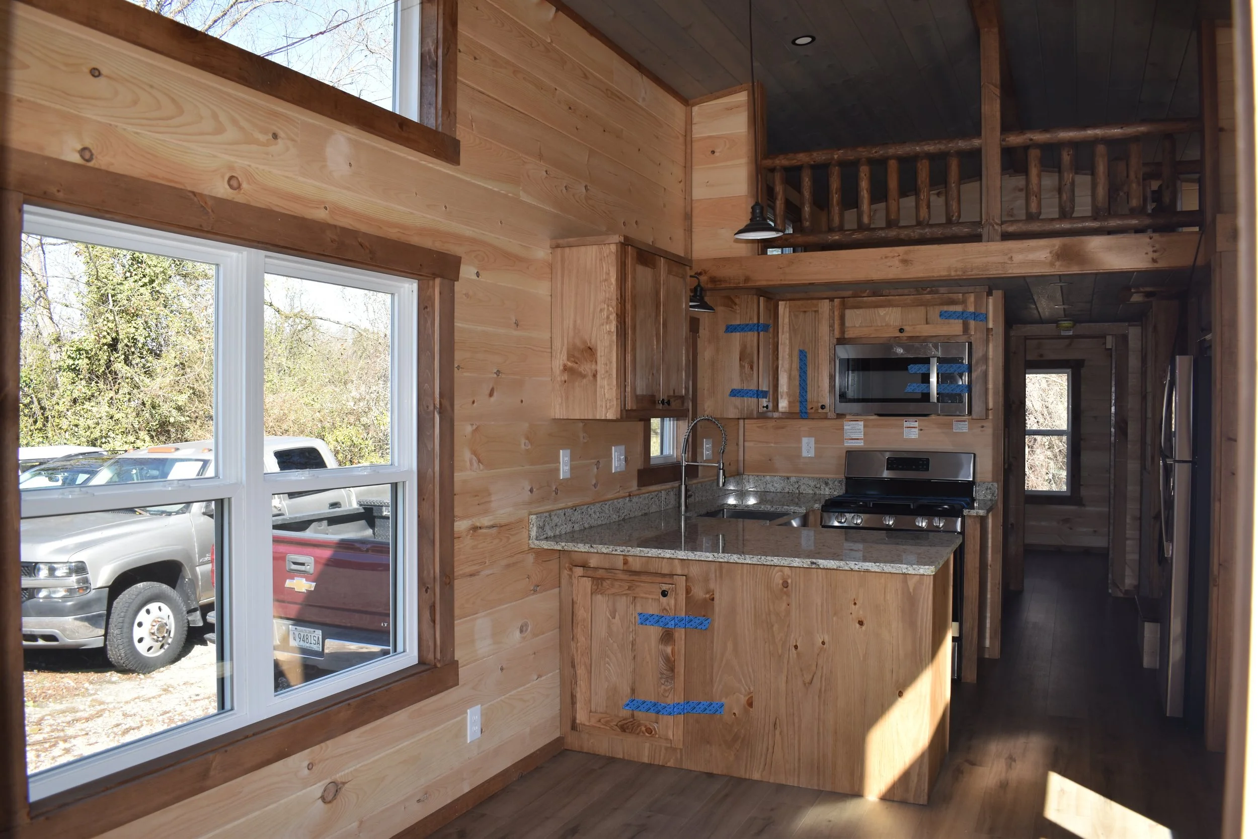 Interior of a wooden kitchen with granite countertop, stainless steel microwave and stove, and large window showing outdoors parking lot with white and gray trucks.