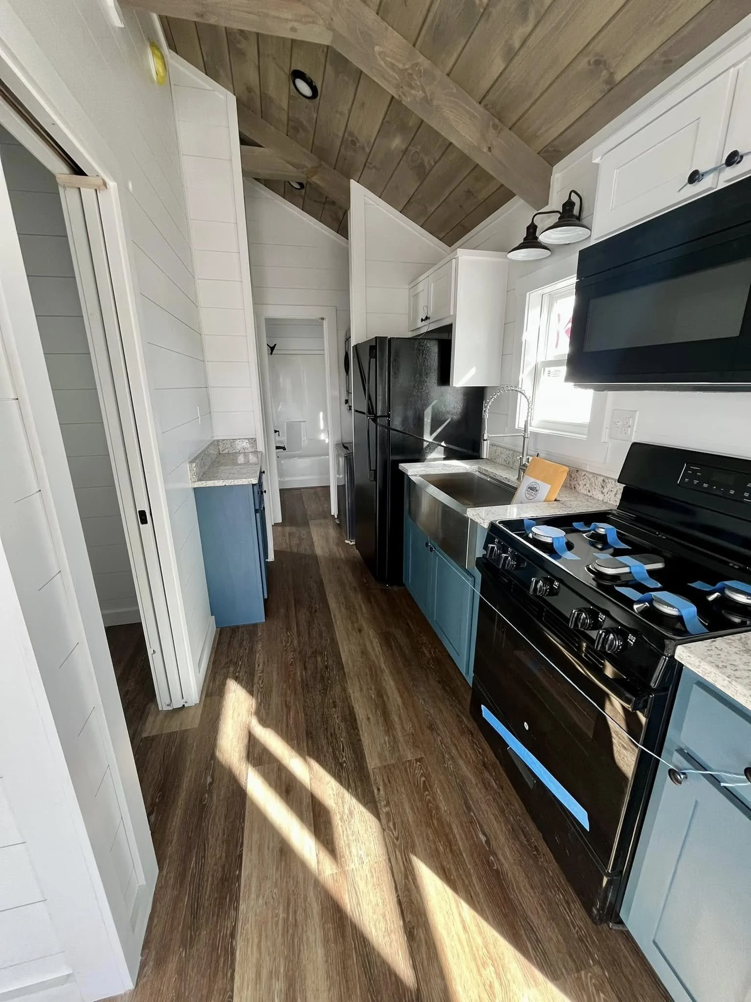 A modern kitchen with white shiplap walls, wood ceiling, black appliances including a refrigerator and stove, blue cabinets, a granite countertop, a large stainless steel sink, and a small window letting in natural light.