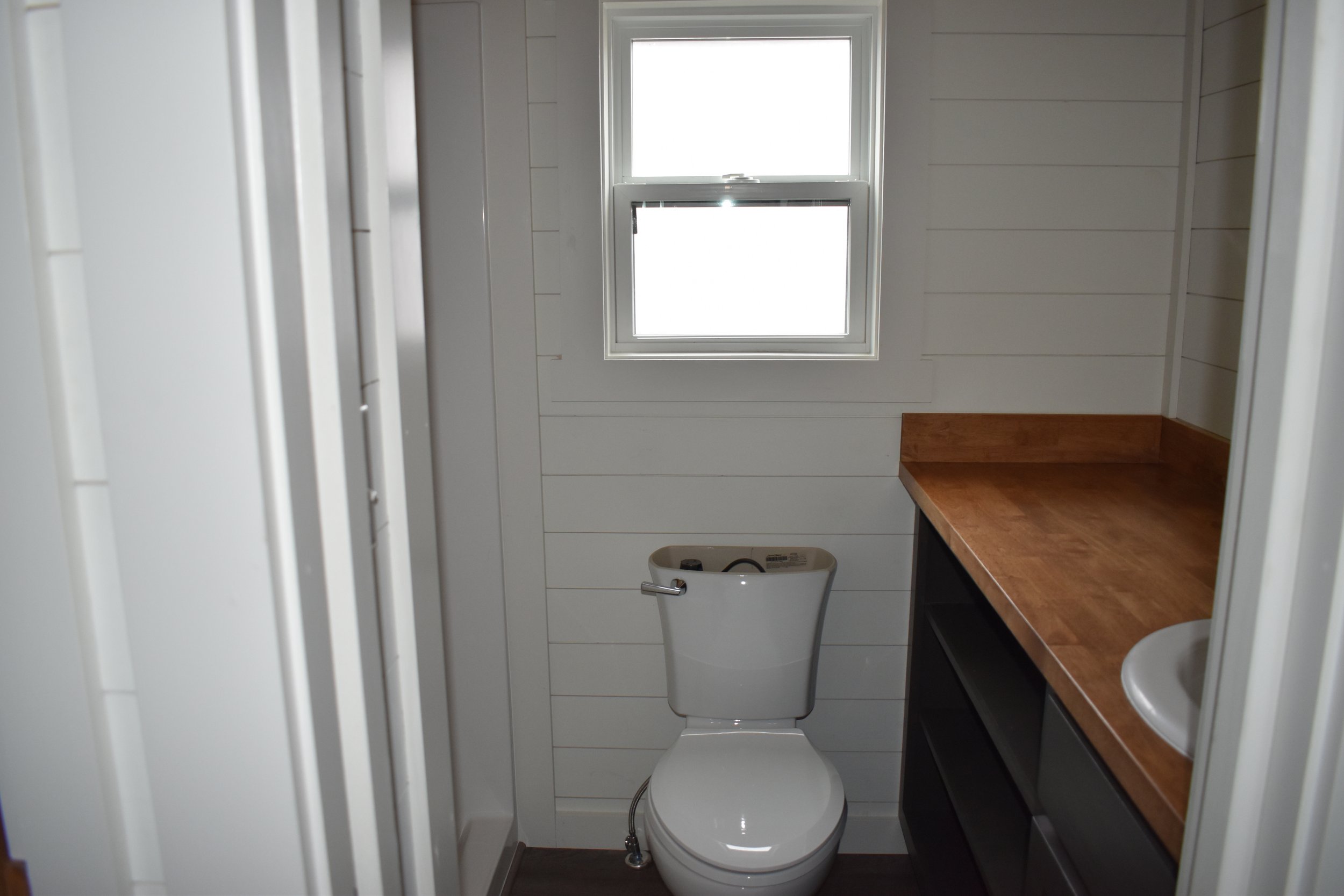 A small bathroom with white shiplap walls, a wooden countertop with a white sink, a window, and a white toilet.