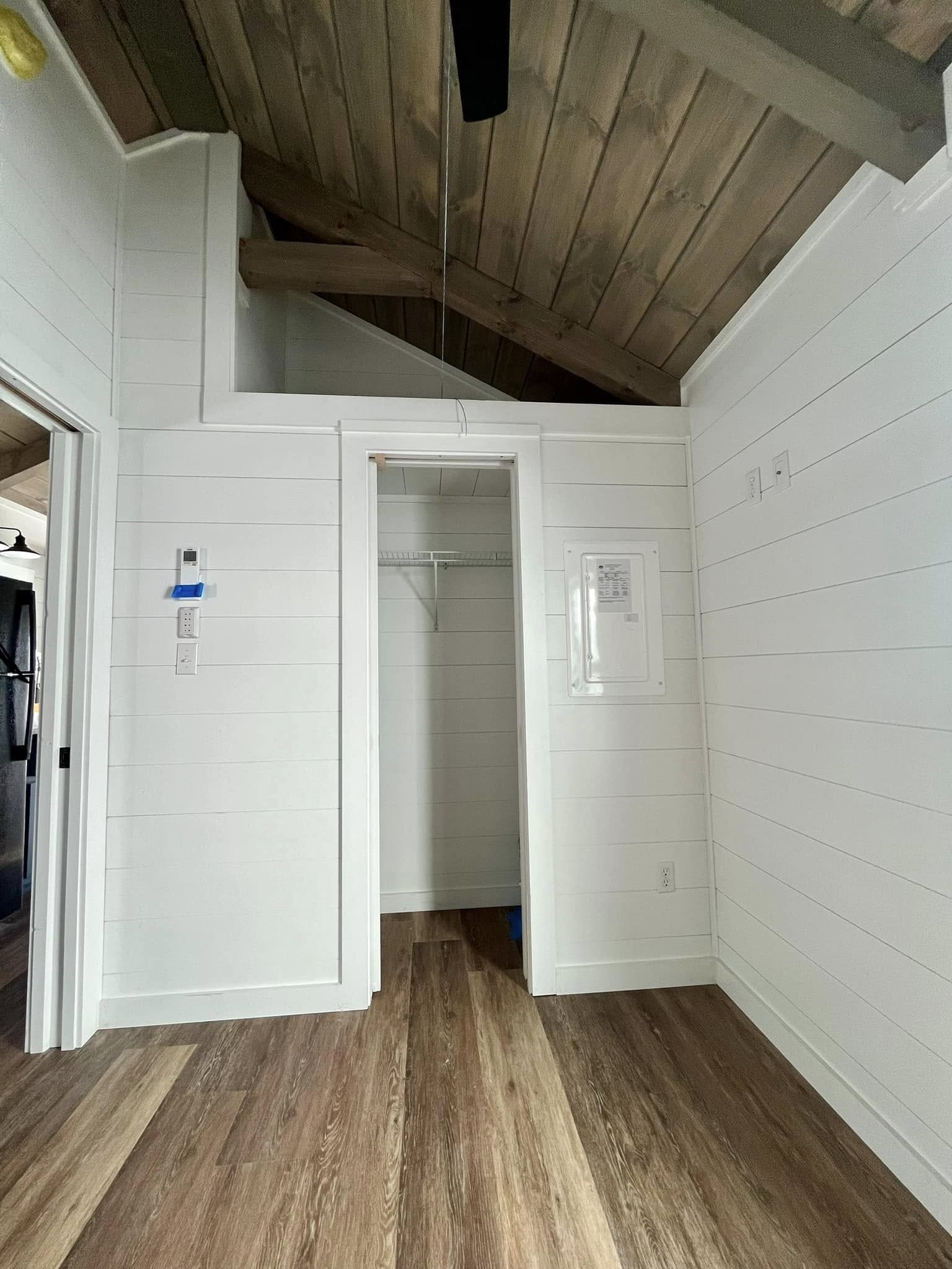 Empty closet with open door, white shiplap walls, and wood flooring, with exposed wooden ceiling beams.