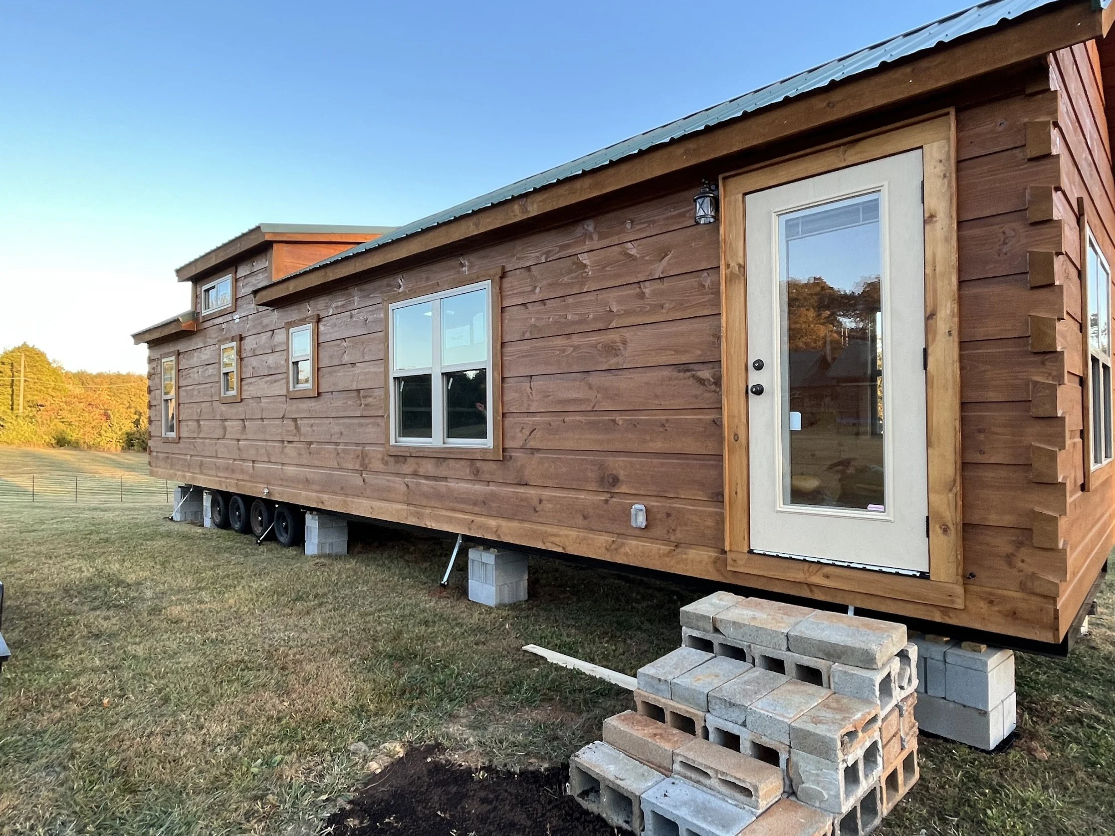 A wooden tiny house on wheels elevated on concrete blocks with a stack of bricks underneath. The house has multiple windows and a door with a glass panel, a metal roof, and outdoor lighting. It is situated on a grassy field with trees in the background under a clear blue sky.