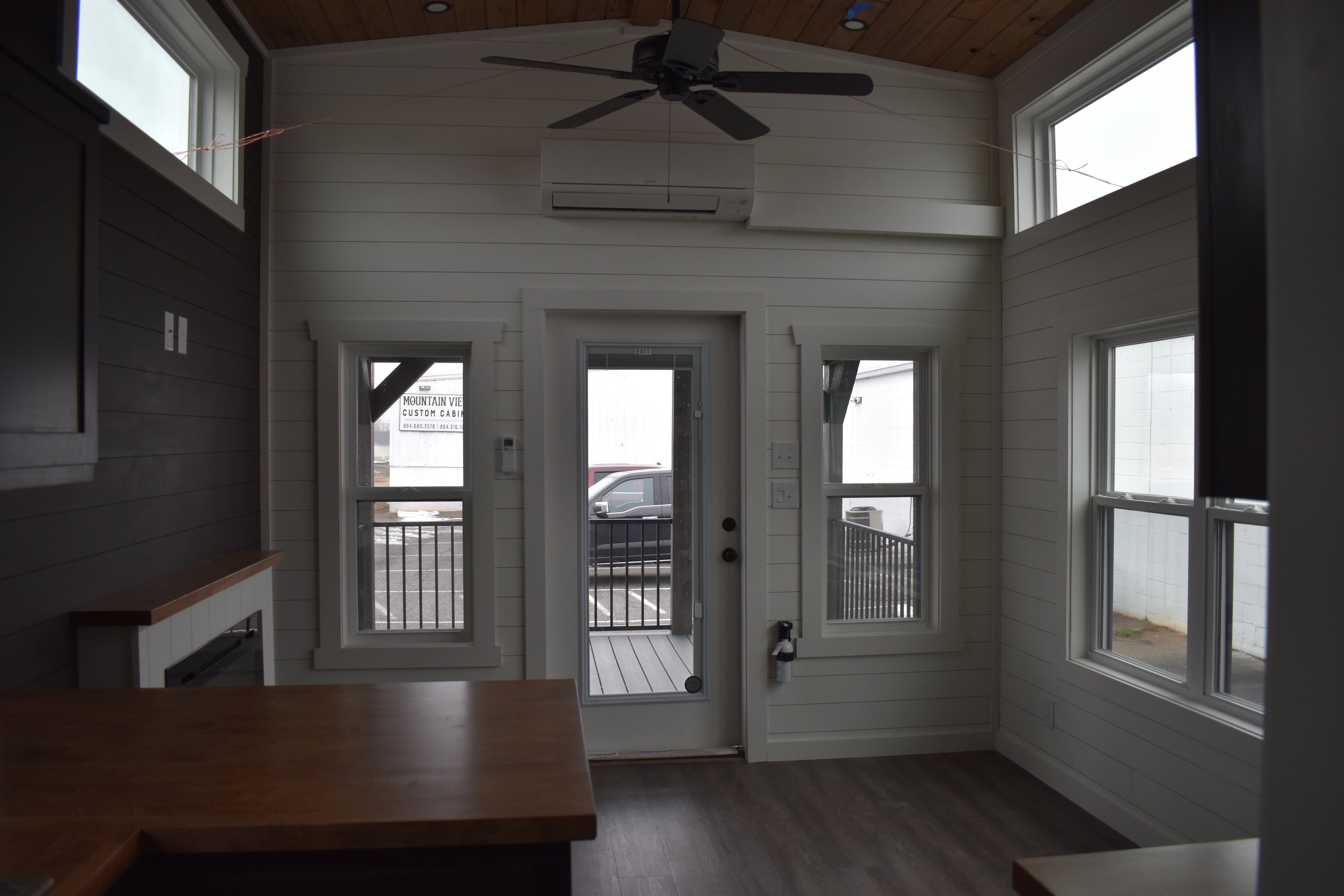 Interior view of a small room with white wooden panel walls, three large windows, a ceiling fan, an air conditioning unit, and a white door leading to an outdoor balcony with a black railing.