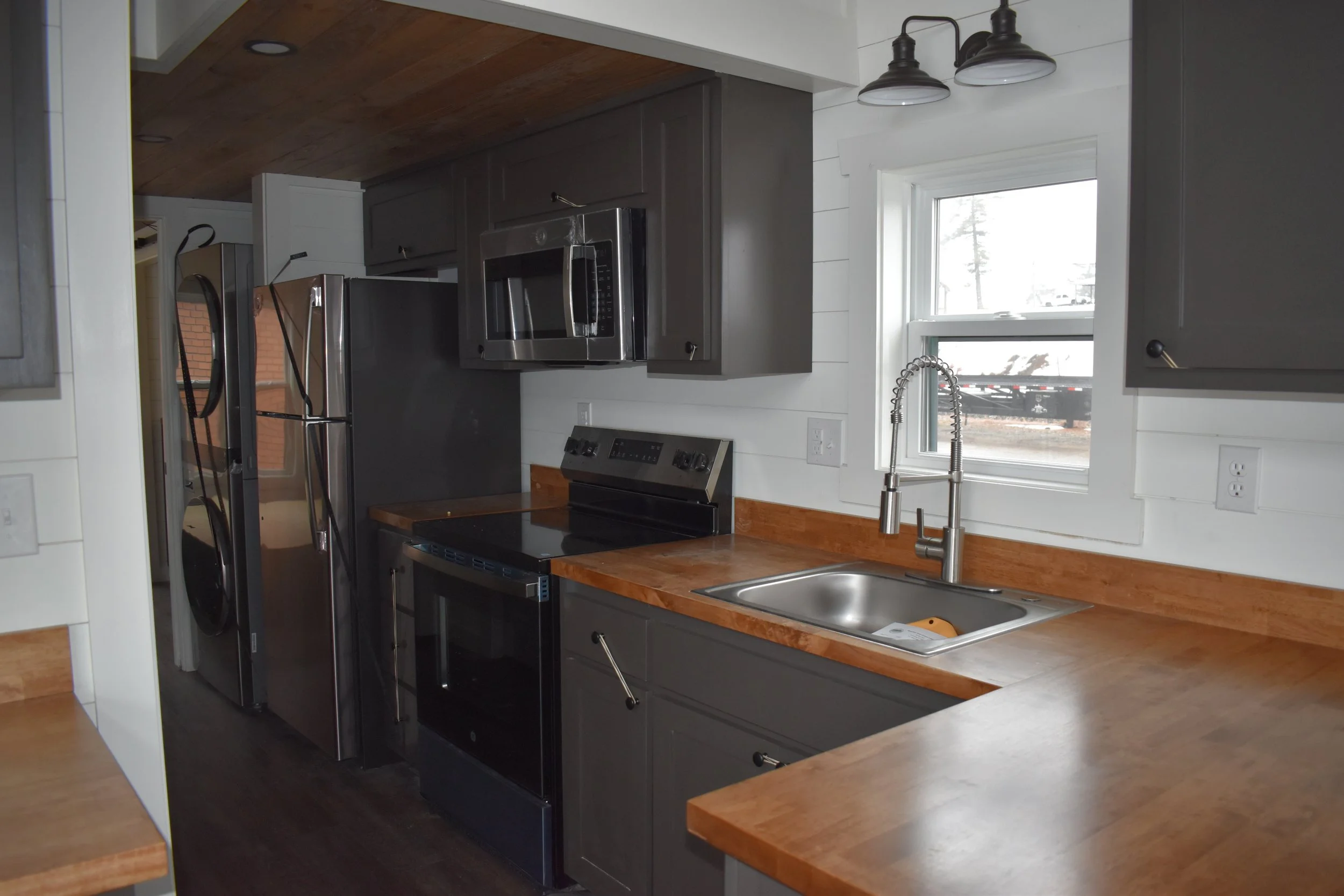 Kitchen with gray cabinets, wooden countertops, stainless steel appliances including microwave, oven, and refrigerator, and a window above the sink with a chrome faucet.