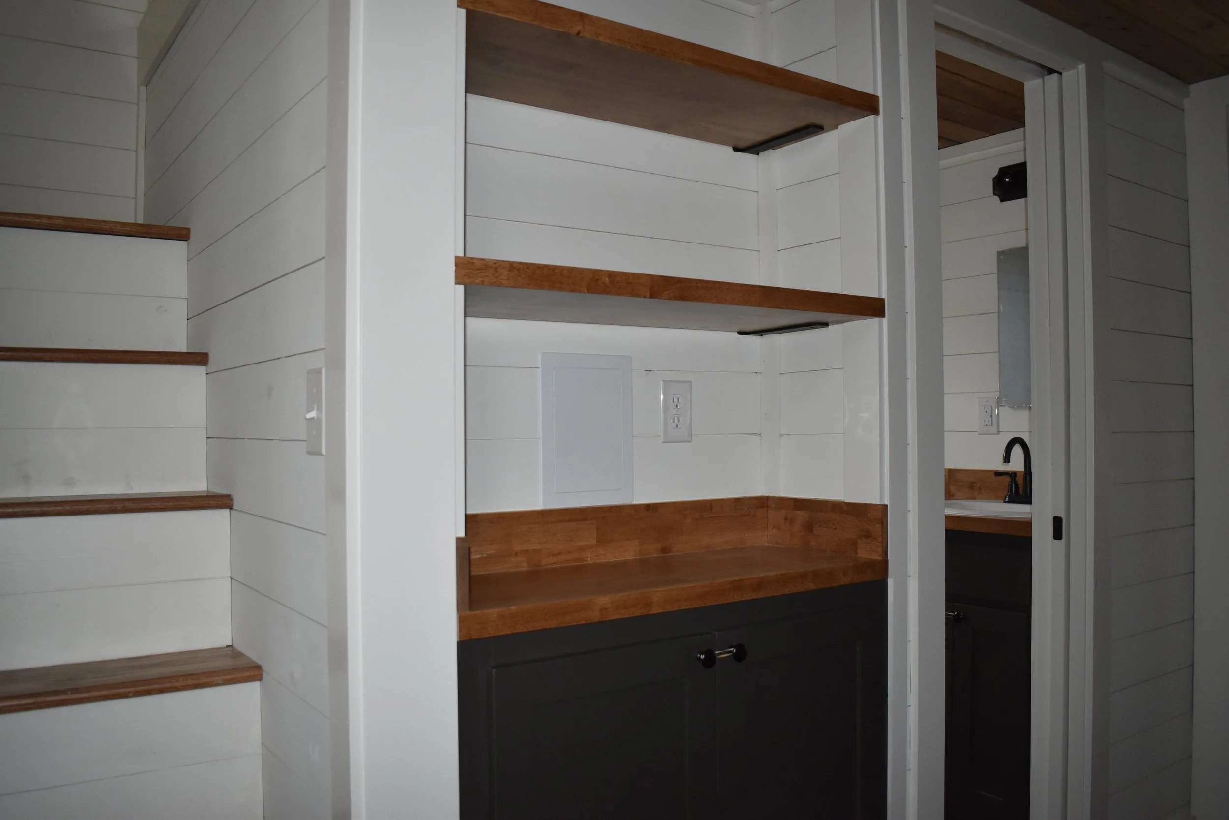 Interior view of a small space with a shelving unit with wooden shelves, a countertop, and a small sink with a black faucet and a white wall panel.