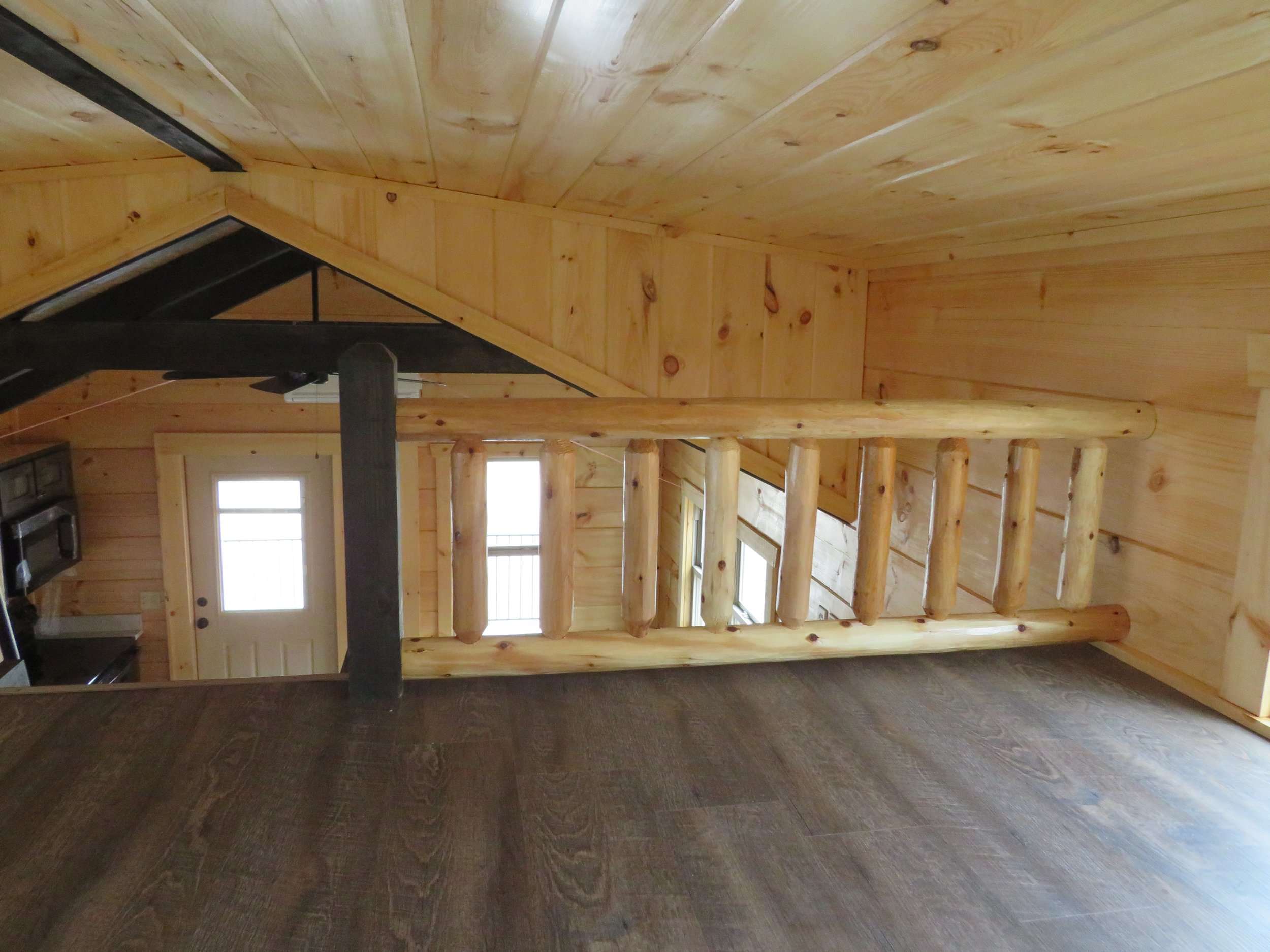 Interior of a wooden house with a sloped ceiling, new wooden flooring, and a staircase with wooden balusters leading down to a door and windows.
