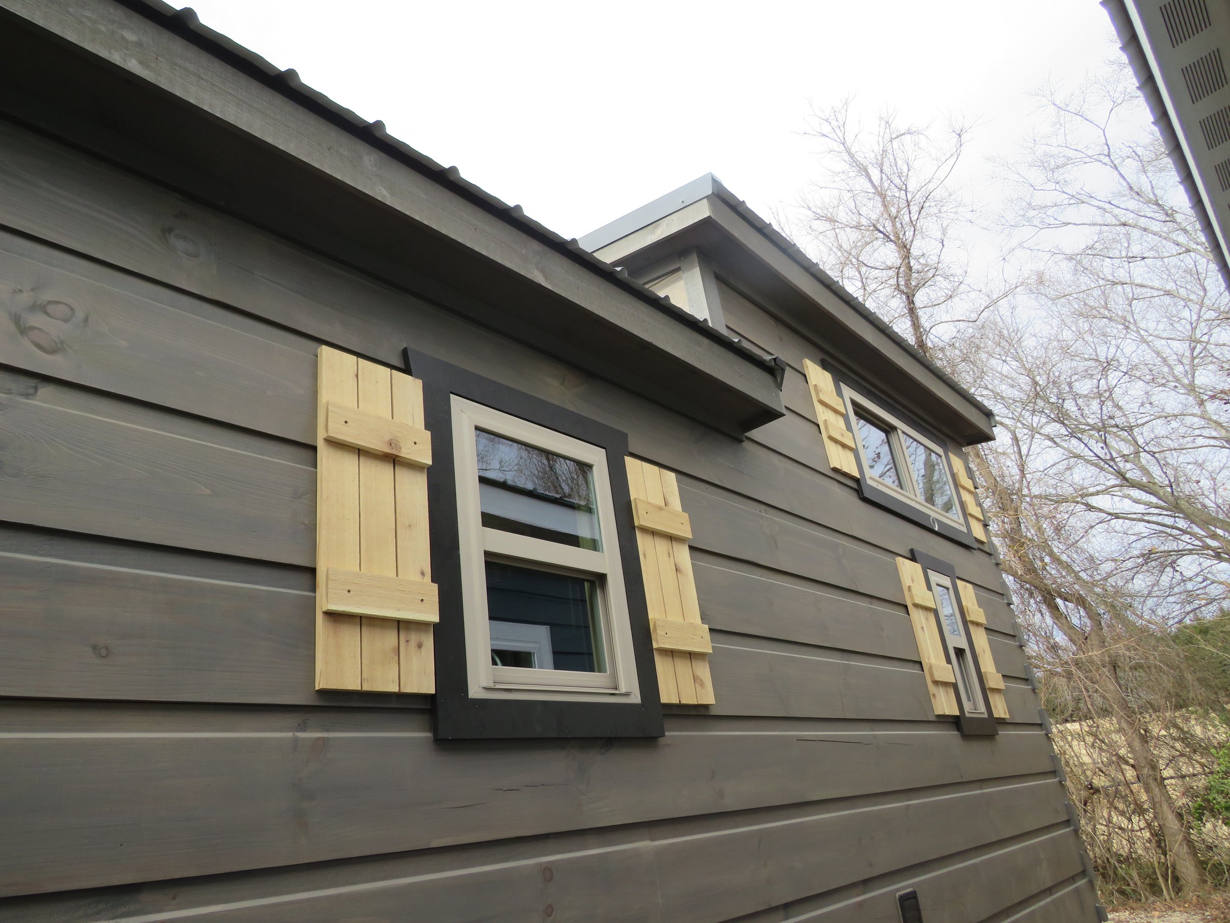 Side view of a house with gray siding, multiple windows with black and gray frames, and light wood shutters in process of being installed.