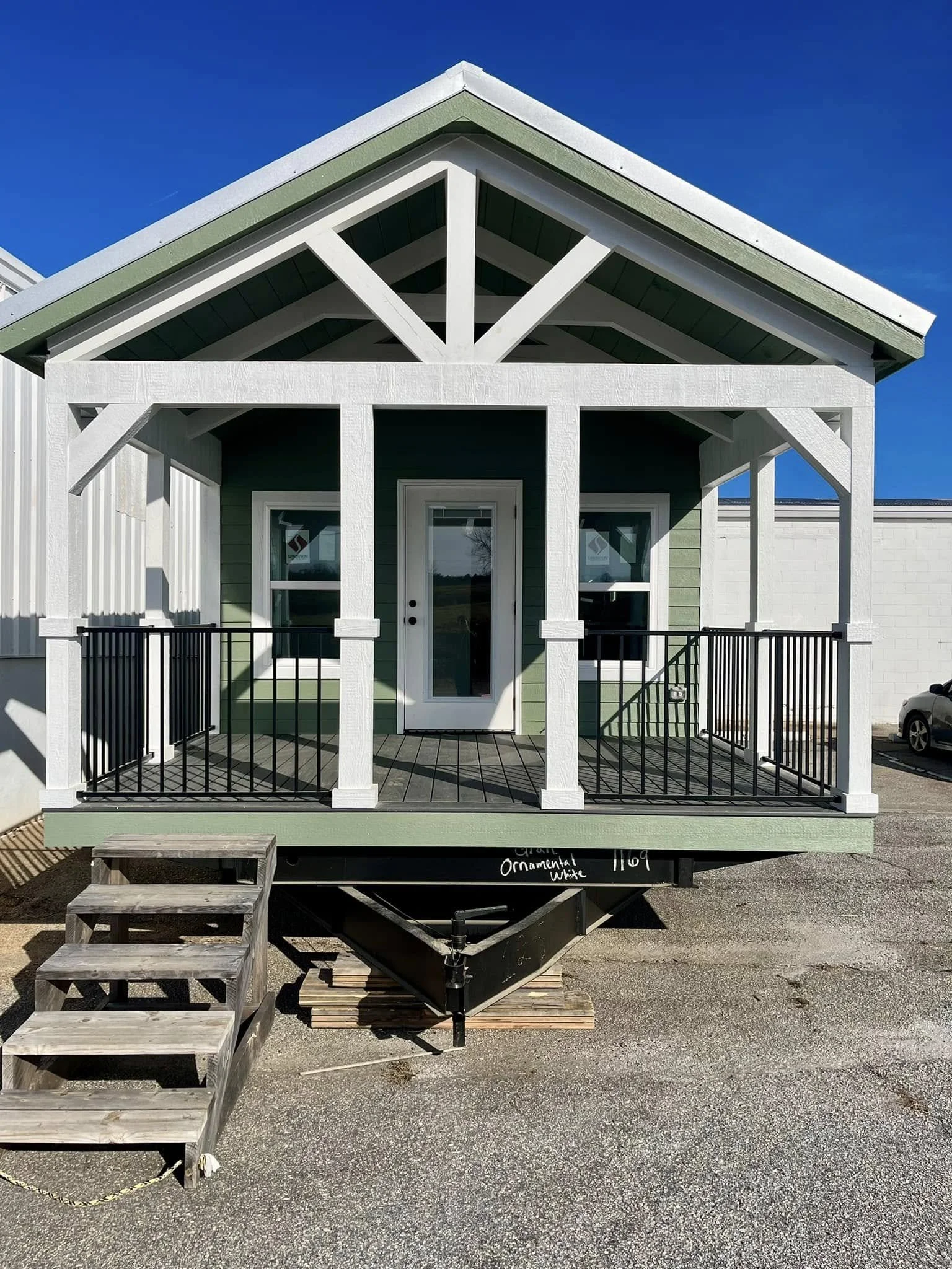 Front view of a small house with a porch, green siding, and white trims, on a trailer.
