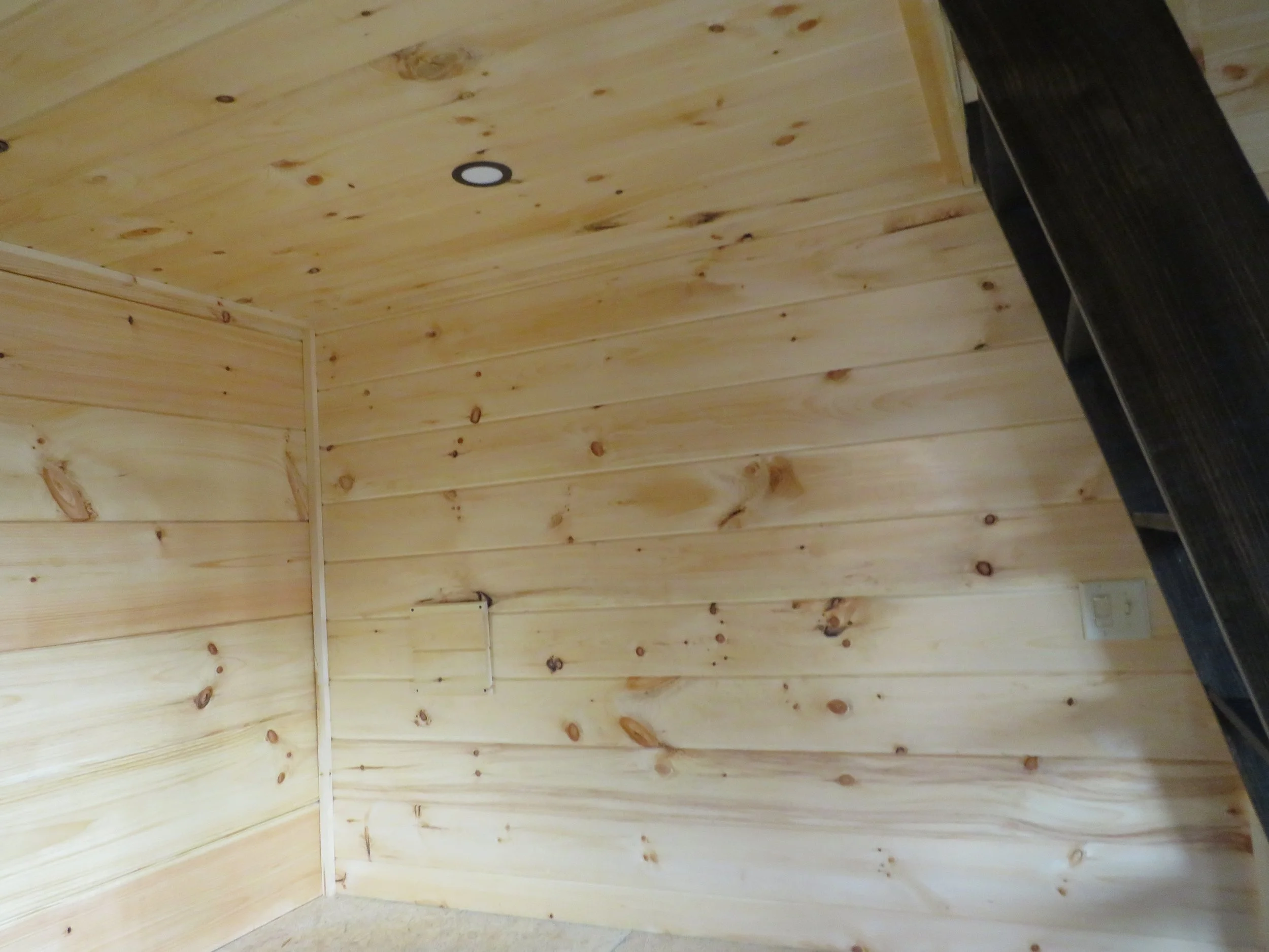 Wood-paneled corner of a room with a small, white electrical outlet and a black staircase or rail on the right side, built with light-colored wooden planks and a ceiling with one recessed light.