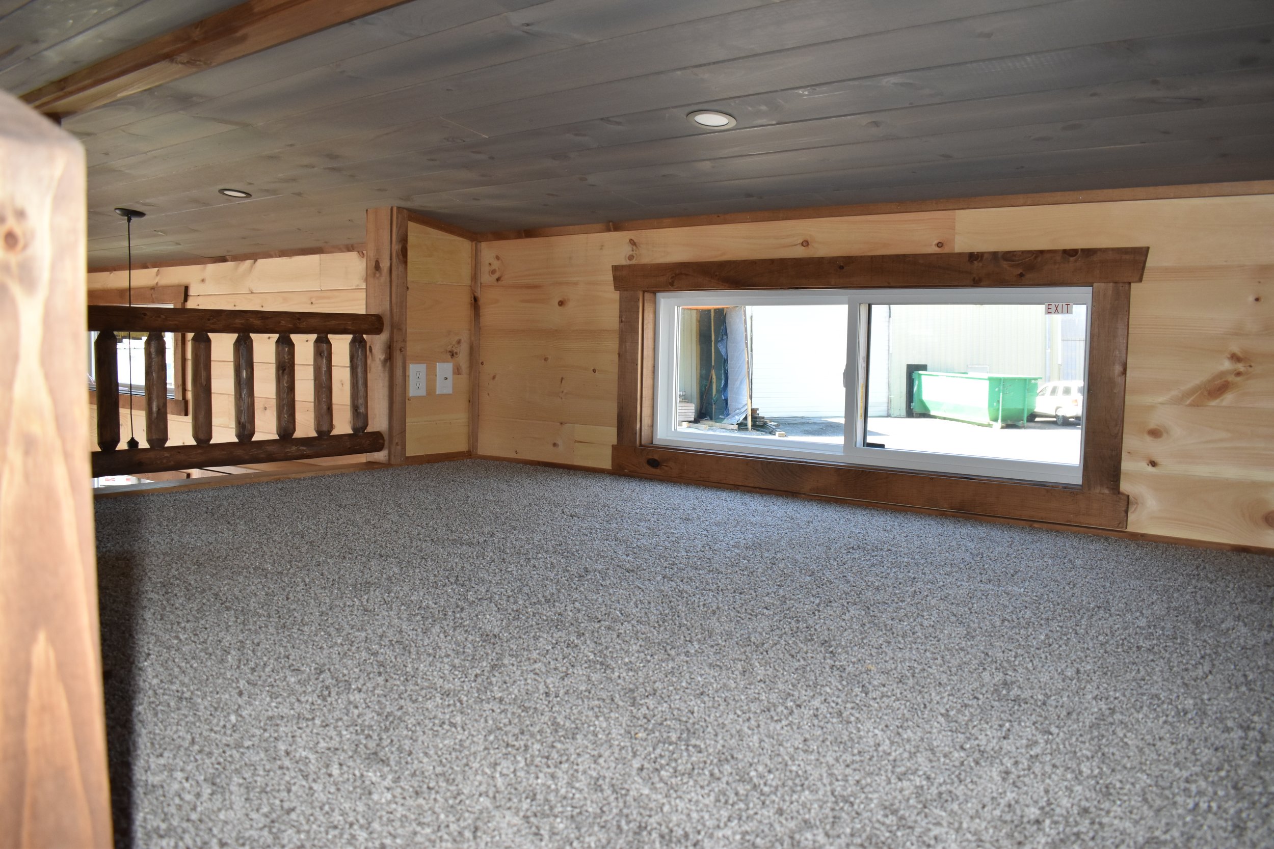 A cozy loft space with wood-paneled walls and ceiling, a large horizontal window, and carpeted floor.