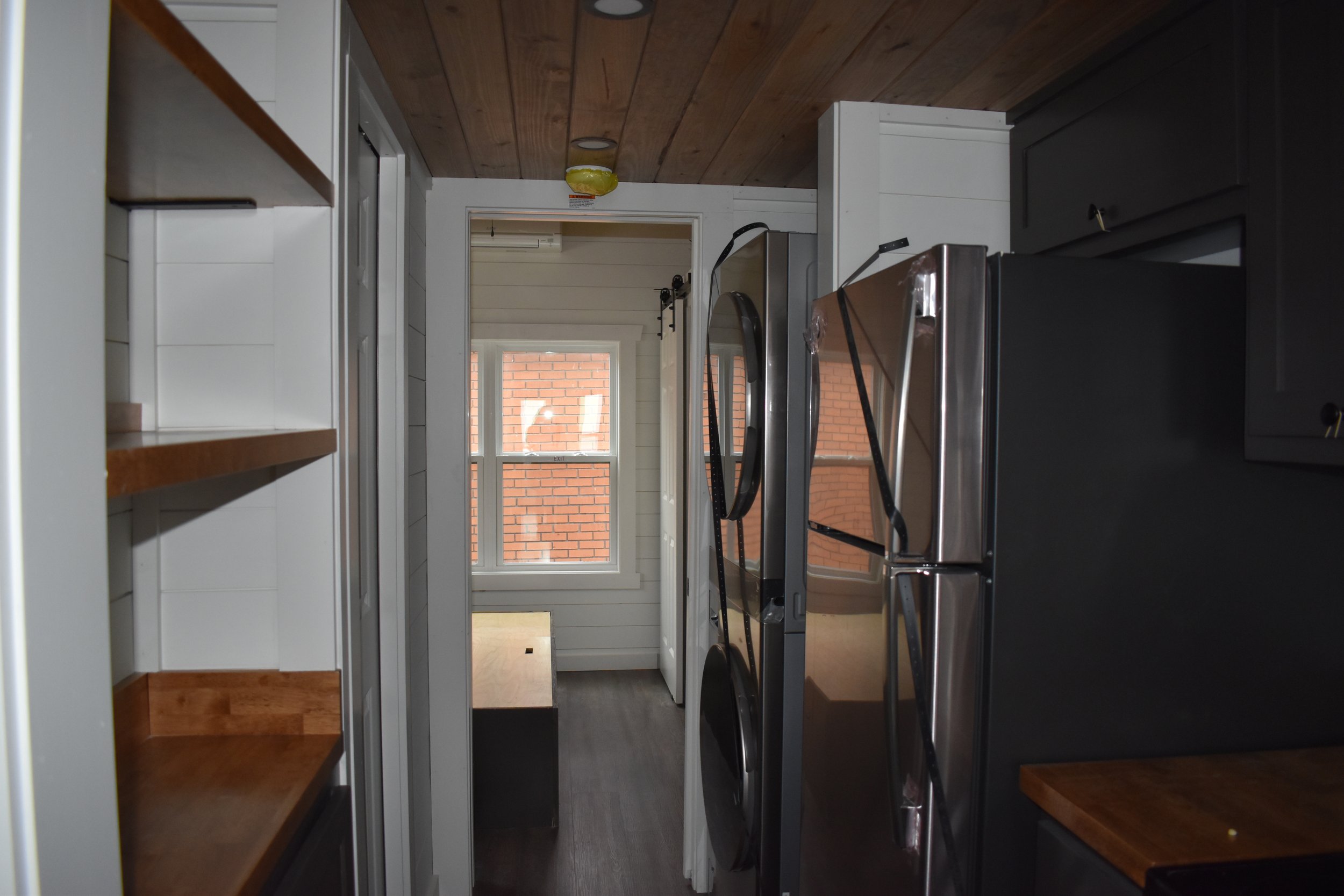 View of a laundry area with a washing machine and dryer, white shelves on the left, gray cabinets on the right, and a window with a brick exterior wall outside.