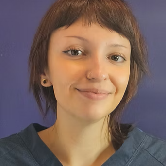 Close-up of a young woman with short brown hair, earrings, and a nose ring, smiling against a purple background.