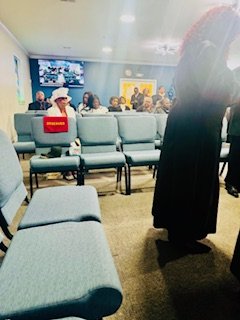 People seated and standing in a waiting room or public space, with gray chairs and bright paintings on the wall.
