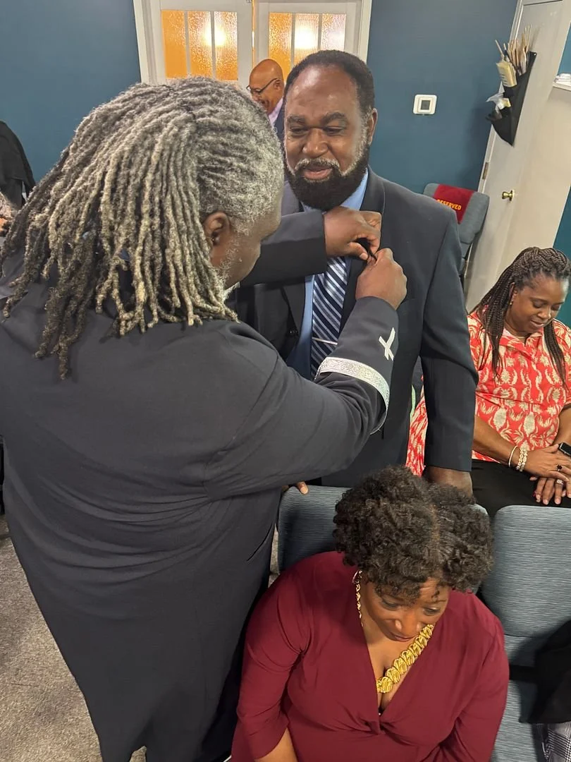 A man in a suit is being awarded a medal by another man with long gray dreadlocks in a black shirt, in what appears to be an indoor ceremony. A woman with short curly hair wearing a maroon dress is seated nearby, smiling and looking down. Two other w