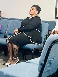 A woman sitting on a blue chair with her eyes closed, wearing a black dress with patterned stockings and beige high heels, appears to be in a waiting or conference room.