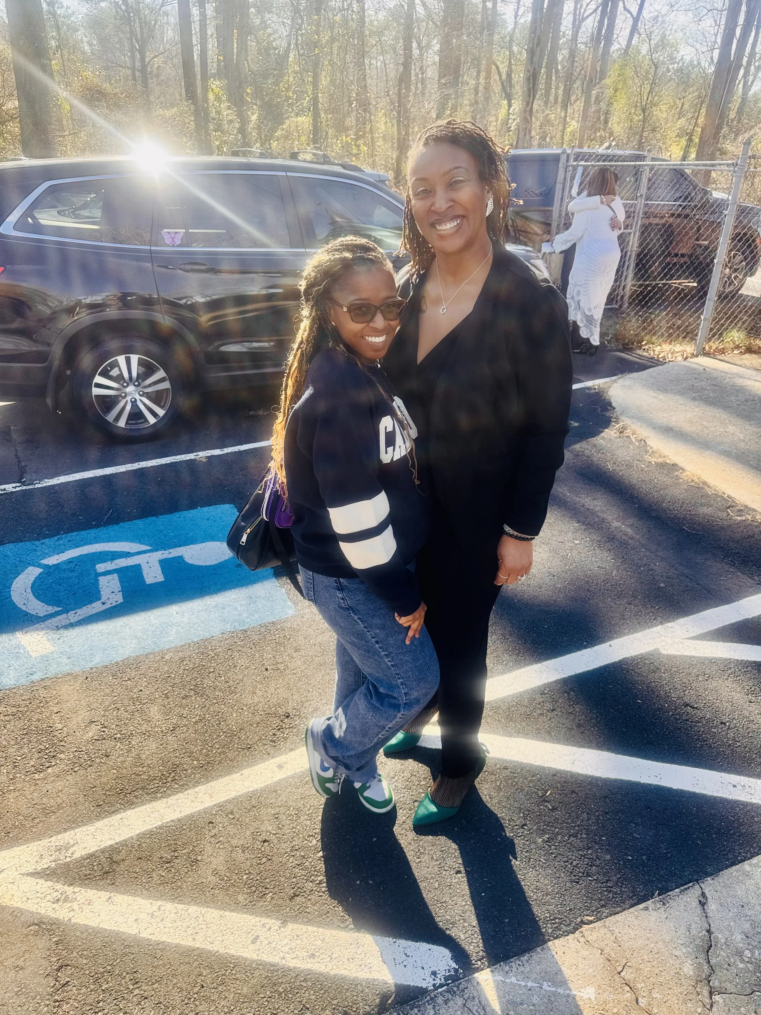 Two women smiling and standing close together in a parking lot with trees in the background, one wearing glasses and a black jacket, and the other with dreadlocks and a black blazer.
