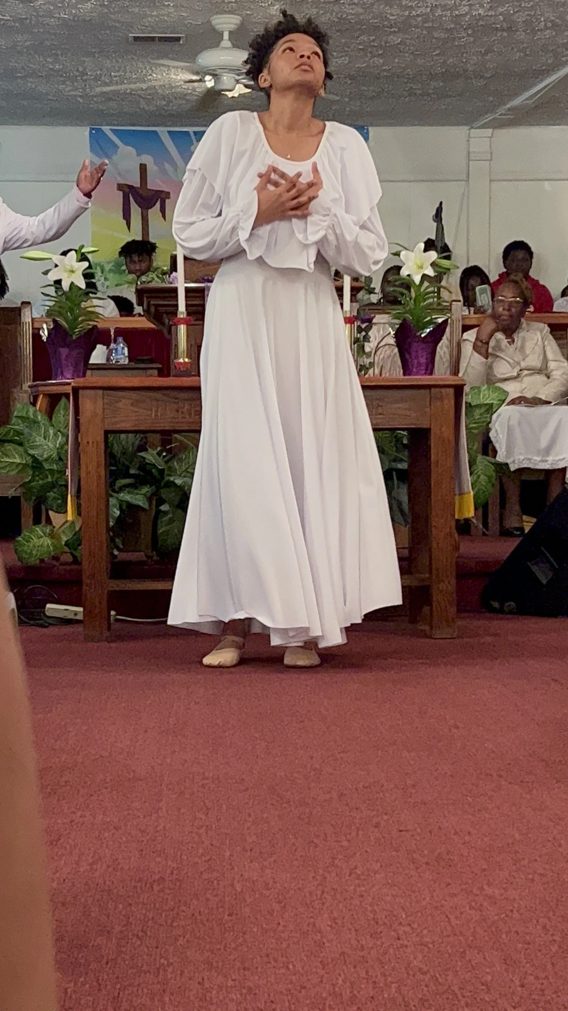 A woman dressed in a long white dress and beige shoes standing with her hands on her chest in a church or religious setting, with other people seated behind her and a cross in the background.