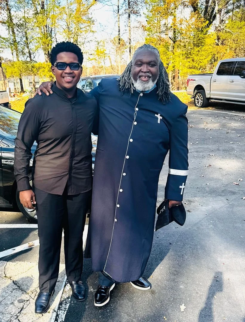 Two men standing outdoors in a parking lot, smiling and posing for the photo, with trees and cars in the background.