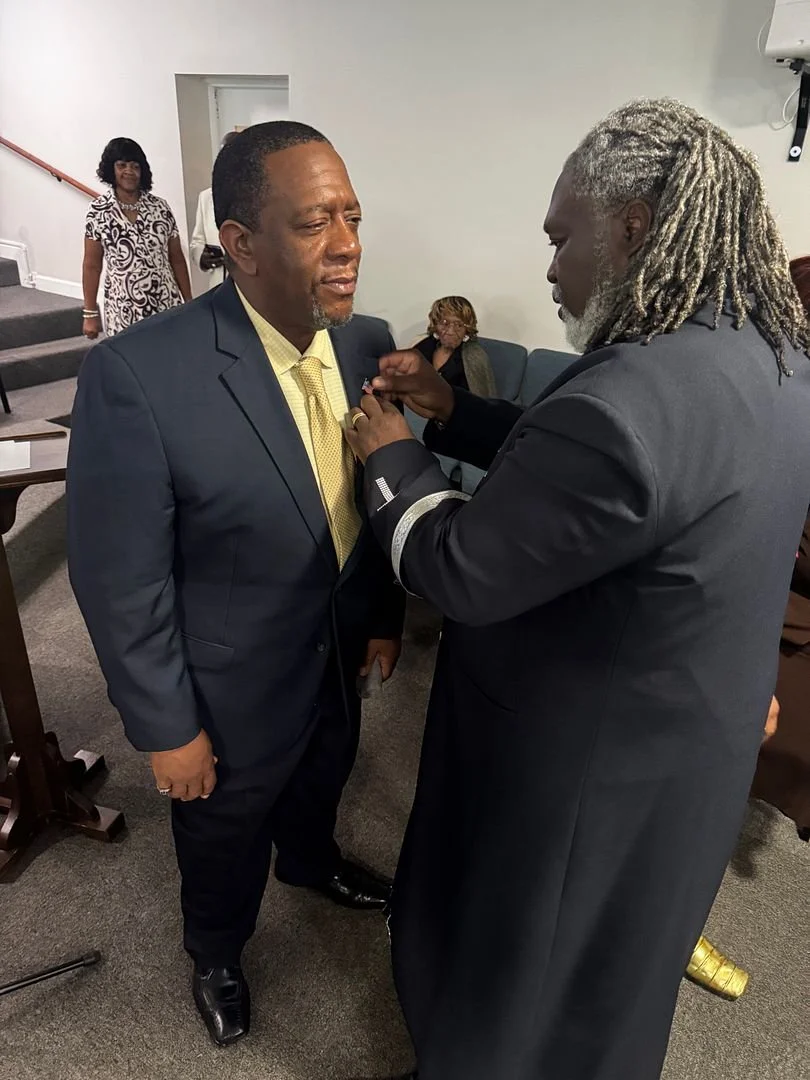 A man in a suit receiving a lapel pin from another man in a suit, with a woman sitting and another woman standing in the background.