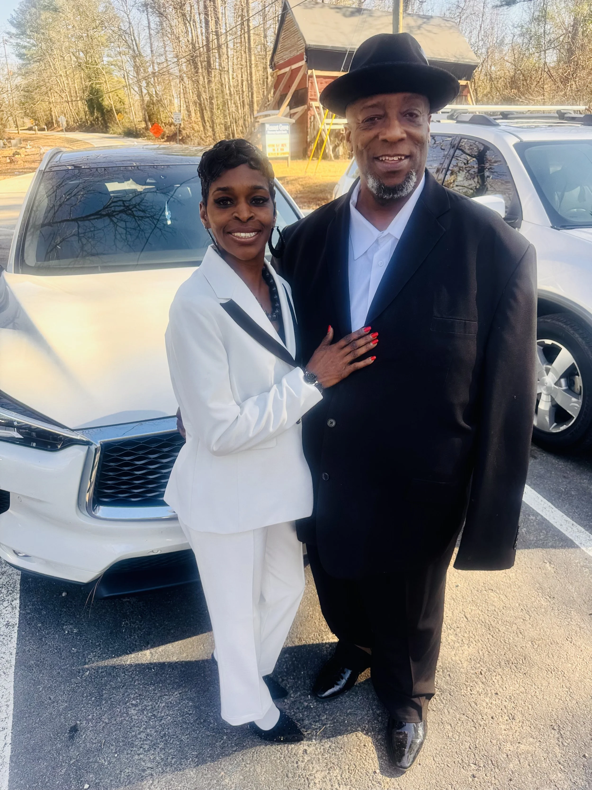 A smiling couple dressed in formal attire standing outdoors in front of parked cars, with a small wooden church and leafless trees in the background.