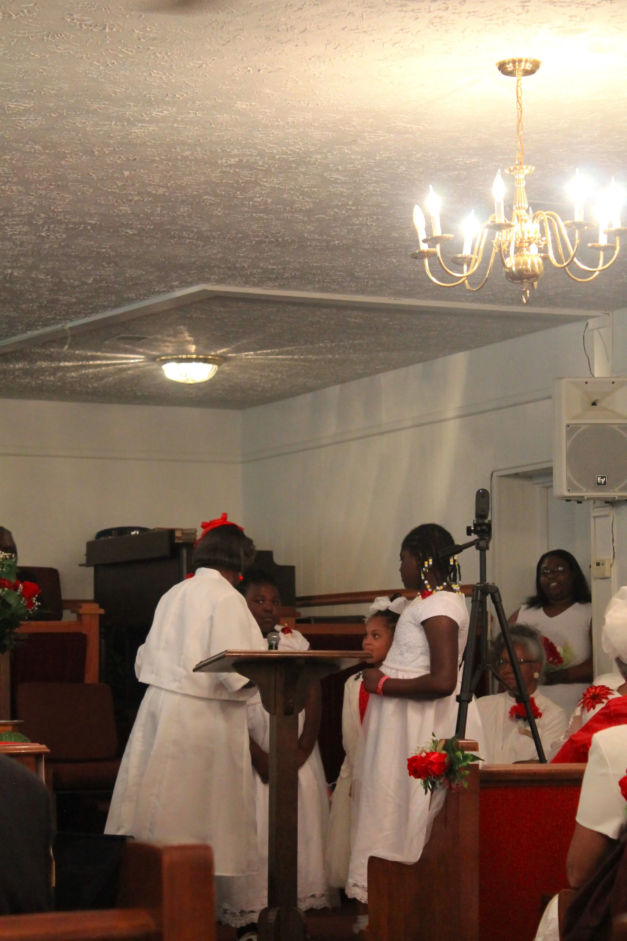 A group of women and children dressed in white clothes attending a church service or ceremony, some holding flowers, with a microphone and camera set up in the room.