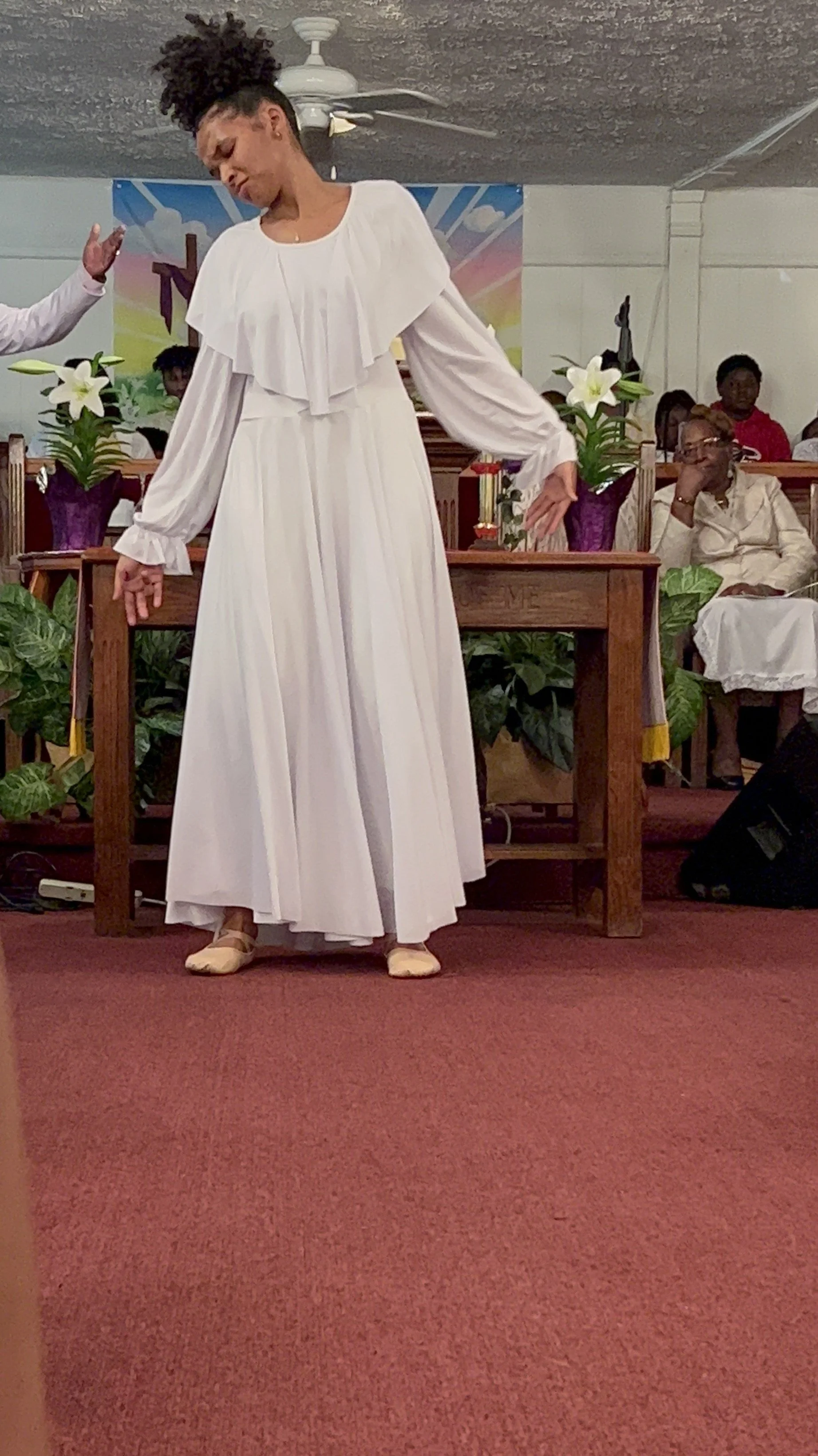 A woman in a white dress standing on a red carpeted floor in a church or religious setting with plants and flowers behind her, and other people sitting in pews in the background.