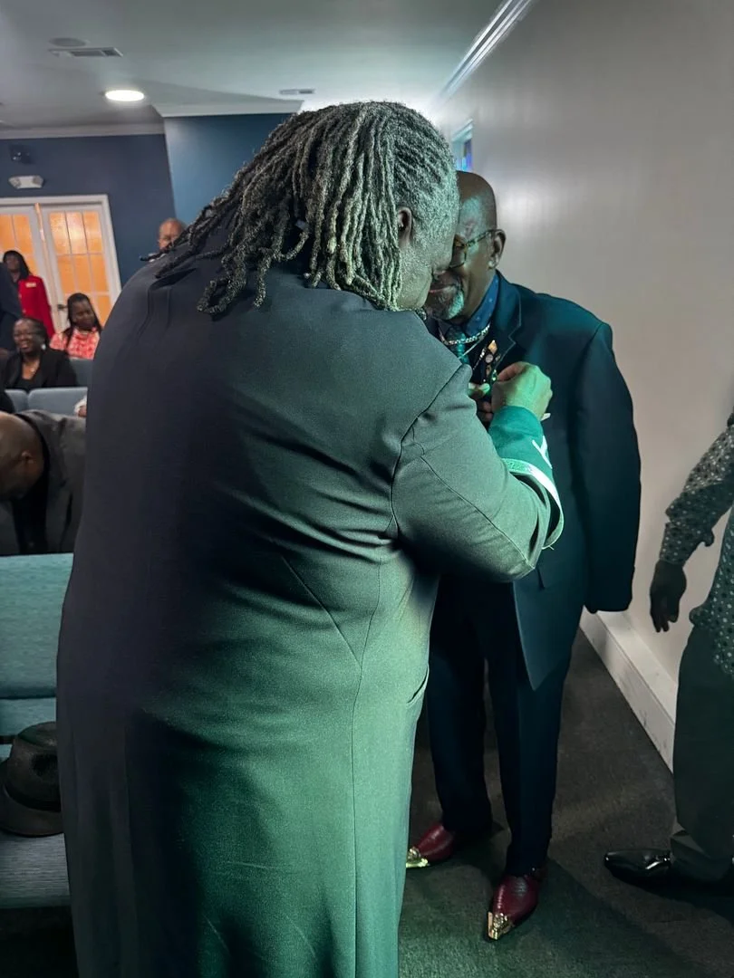 Two men, one with long gray dreadlocks and the other with a beard and glasses, are close together, possibly engaging in a handshake or an embrace, in a room with several onlookers seated in the background.