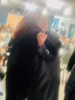 Two people embracing in a crowded indoor setting, possibly a conference or event, with chairs and people in the background.