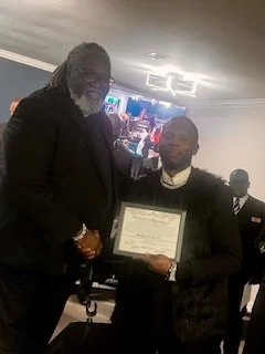 Two men shaking hands, one giving a certificate to the other at an indoor event or awards ceremony.