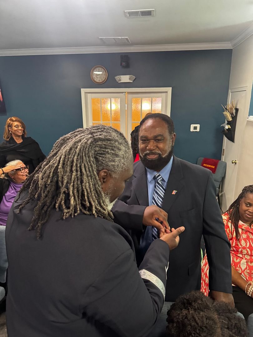 A man in a suit is being pinned with a ring by another man with dreadlocks at a celebration. Several women are watching, smiling in the background.