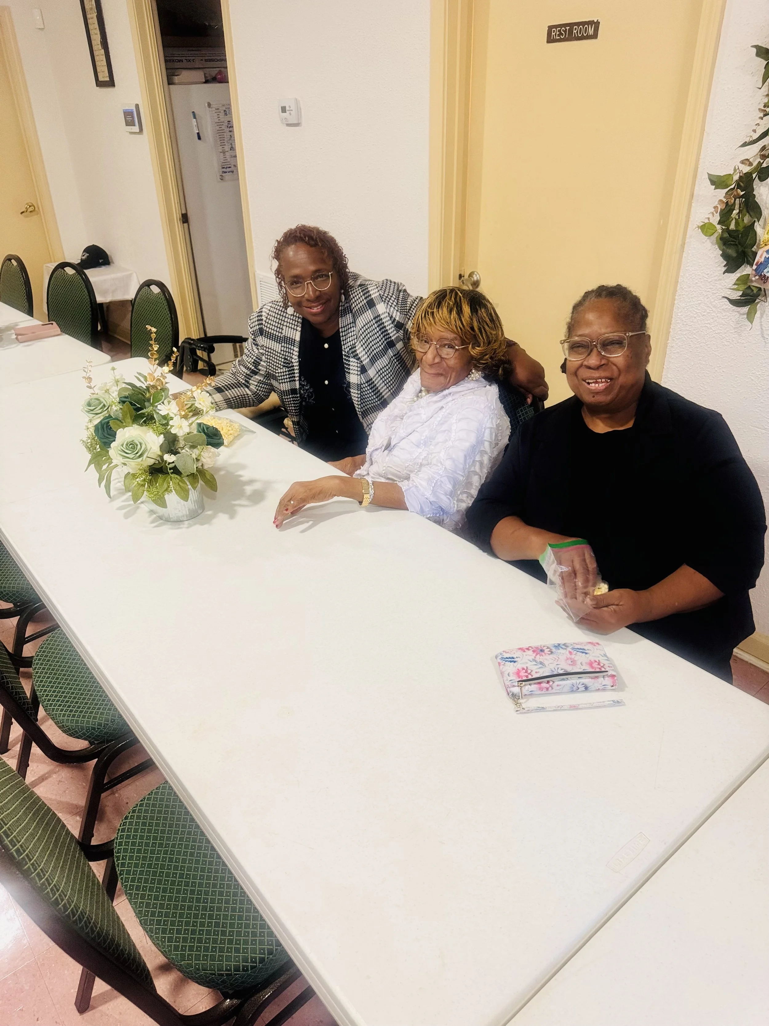 Three women sitting at a long white table with a floral centerpiece, indoor setting, woman on the right holding a small gift, all smiling.