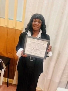 A woman with black hair and glasses holding a framed certificate or diploma, standing indoors in front of a cream-colored curtain.