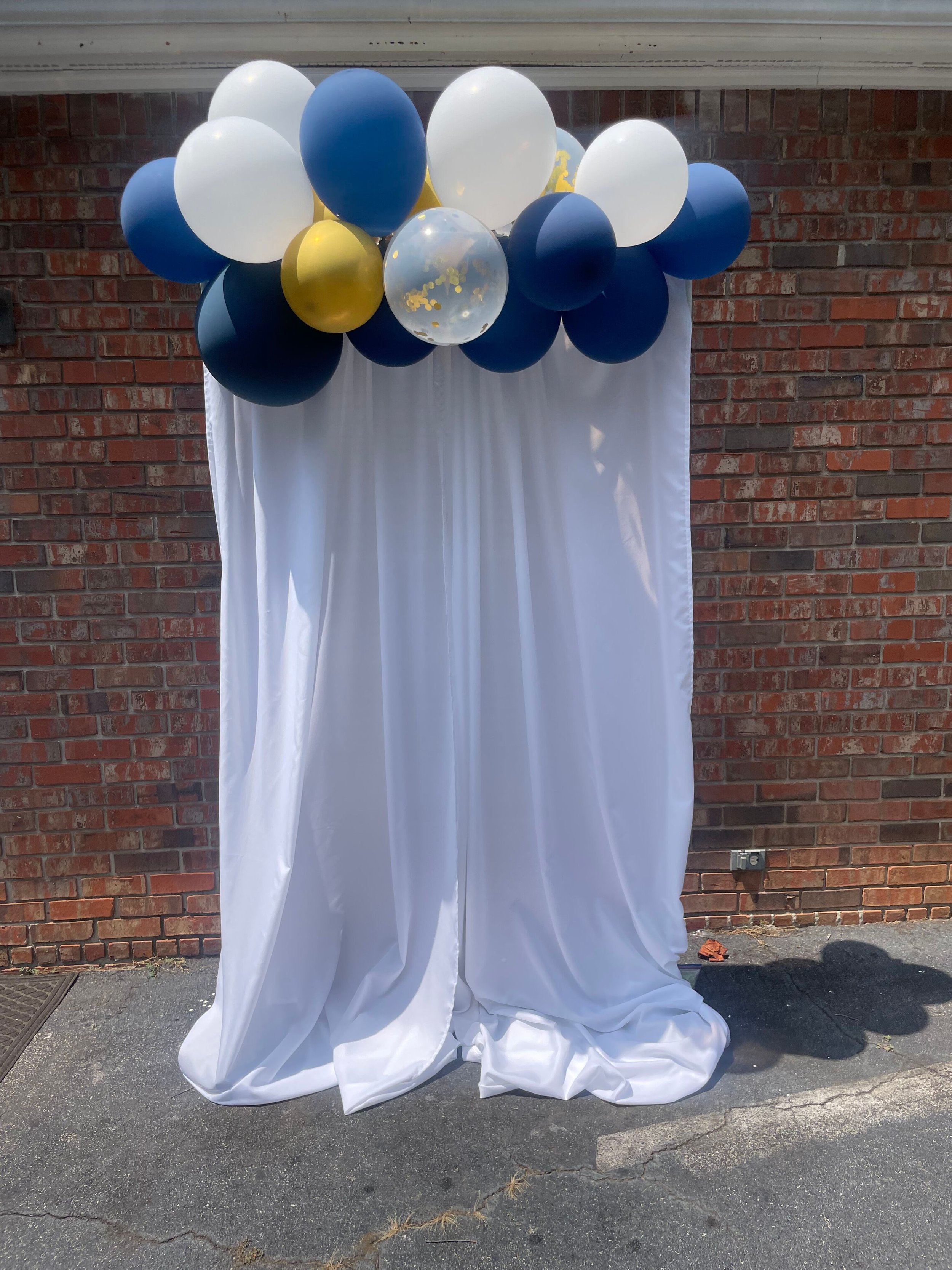 Decorative setup with a bunch of navy blue, white, gold, and clear balloons, some with gold confetti, arranged above a white fabric backdrop against a brick wall.