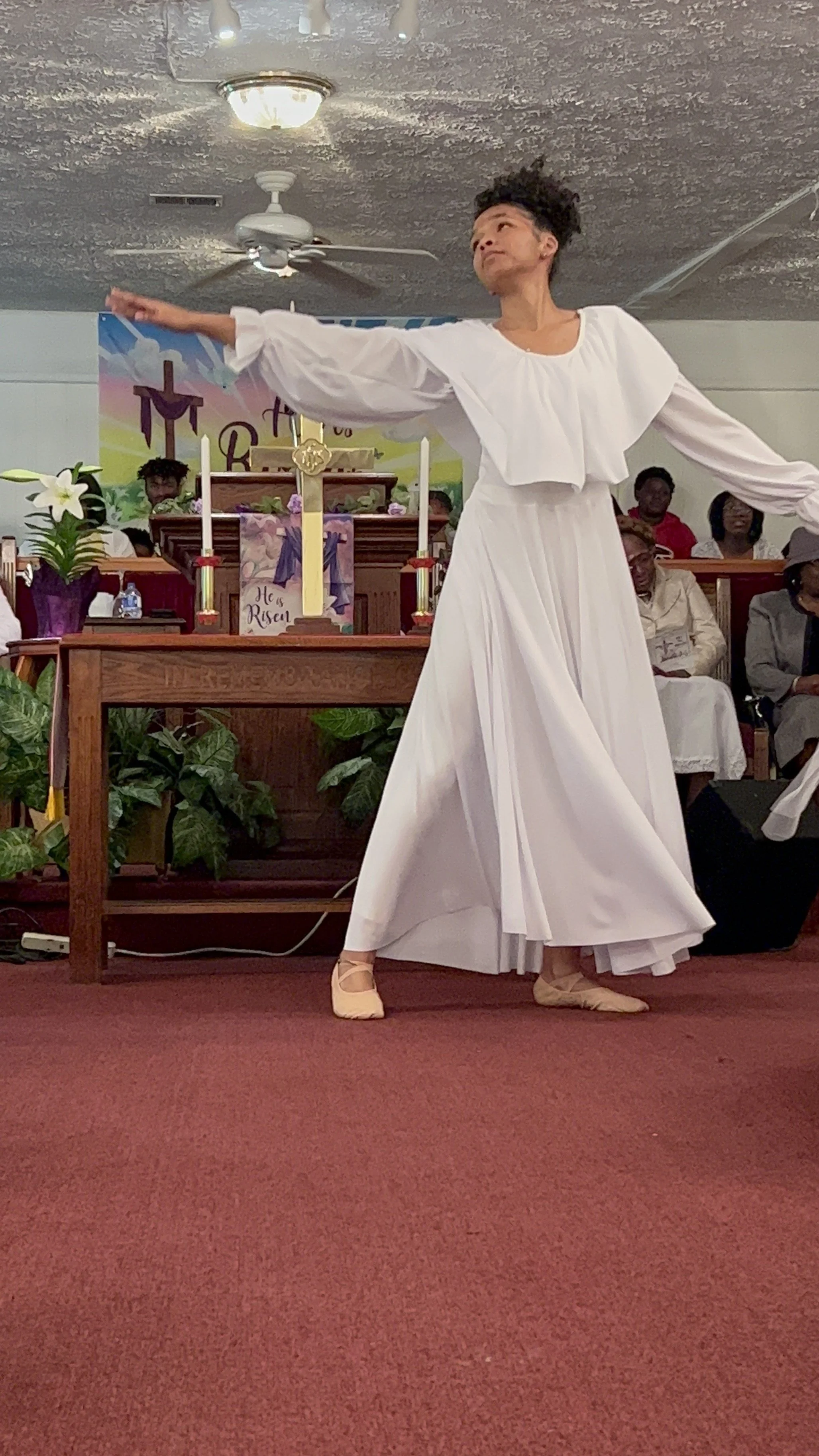 A woman in a white dress dancing in a church, with an audience and church altar in the background.