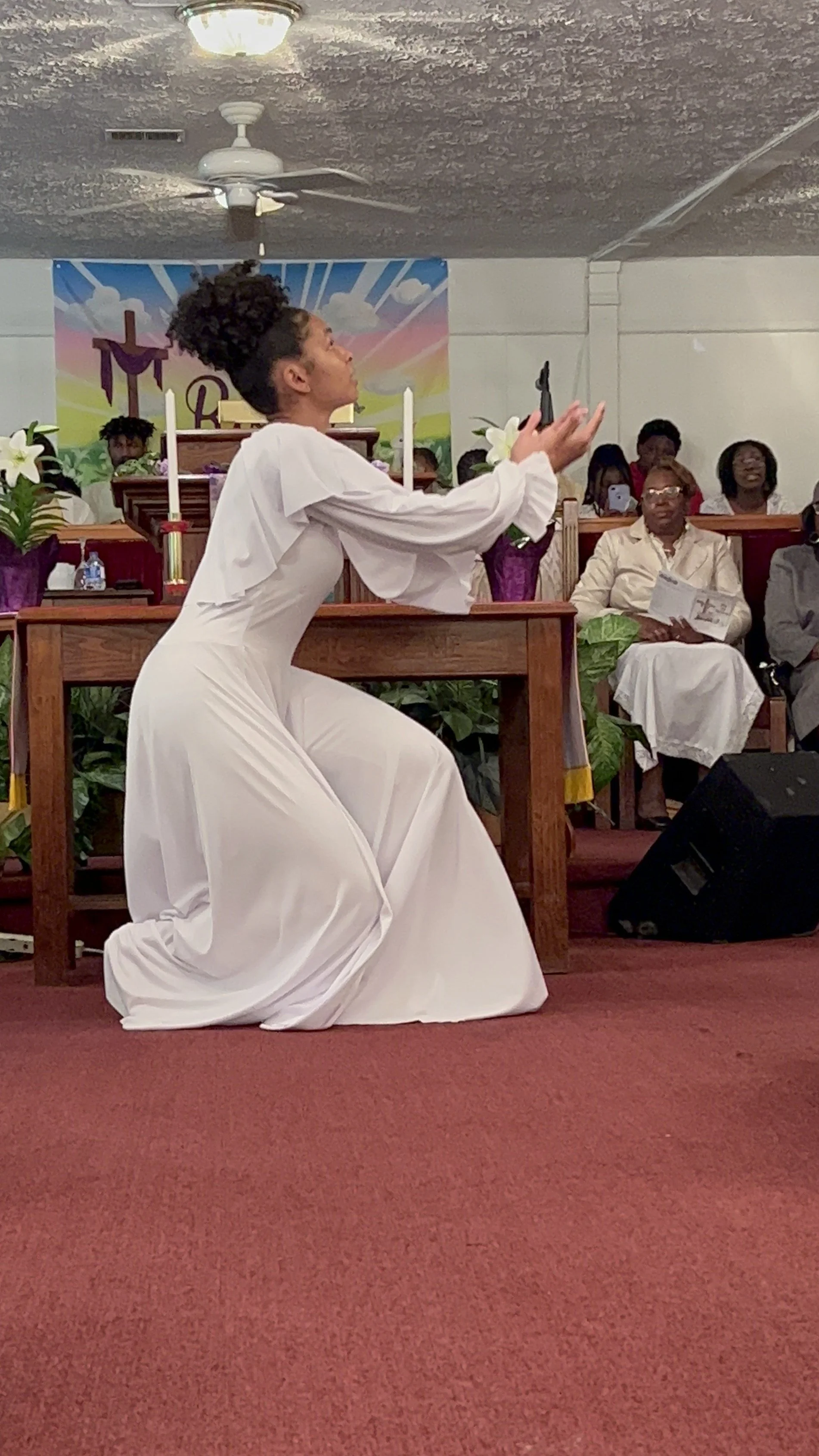 A woman dressed in a white gown kneeling on the floor inside a church. She is holding a microphone and seems to be singing or speaking. The church has a wooden altar with candles and floral decorations, and there are several people seated in the back