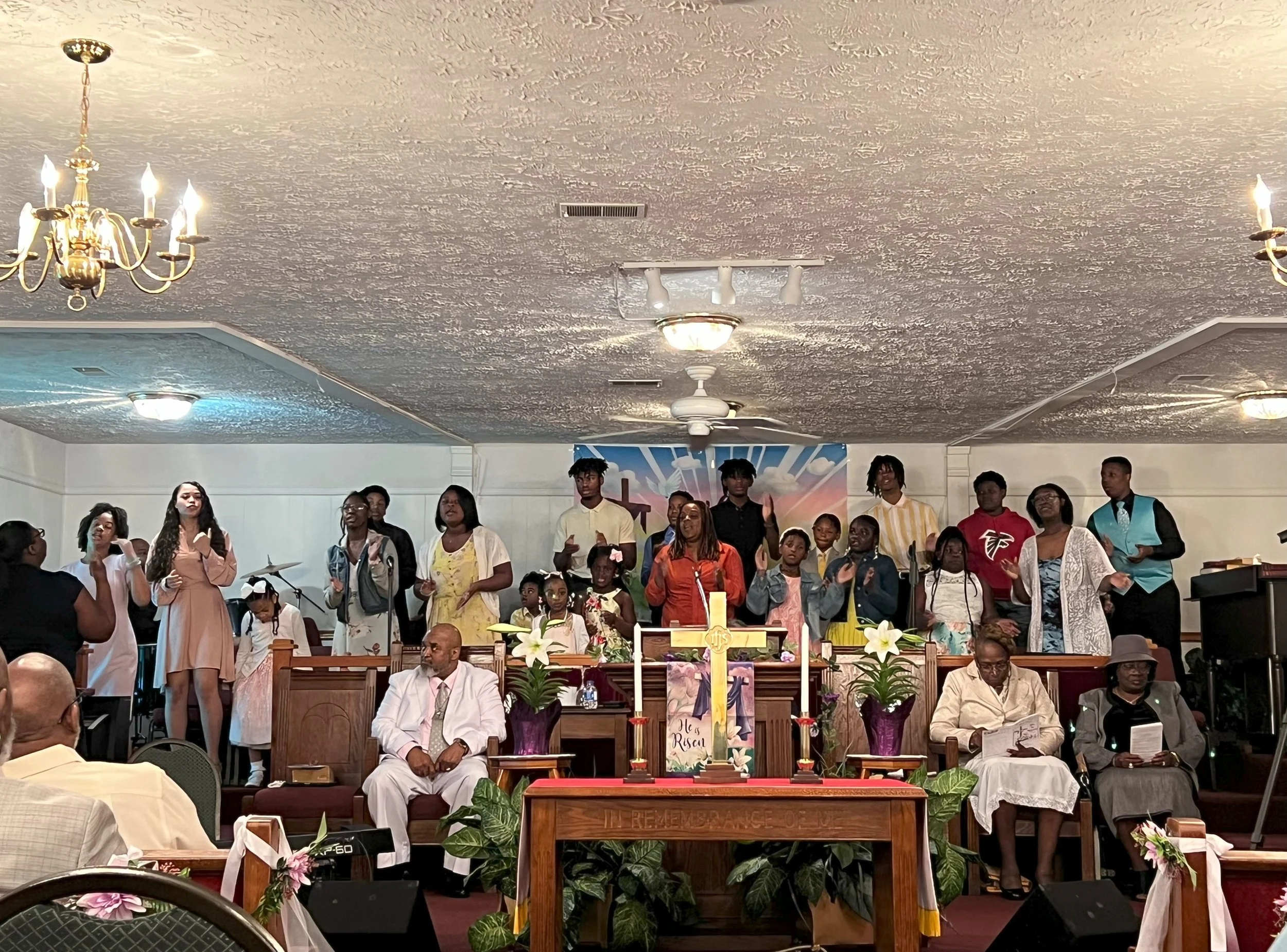 A church service with a diverse group of people, including children and adults, standing on the stage singing; church altar in the foreground with a cross, flowers, and religious items; congregation seated in the pews; ceiling with lights and fans.