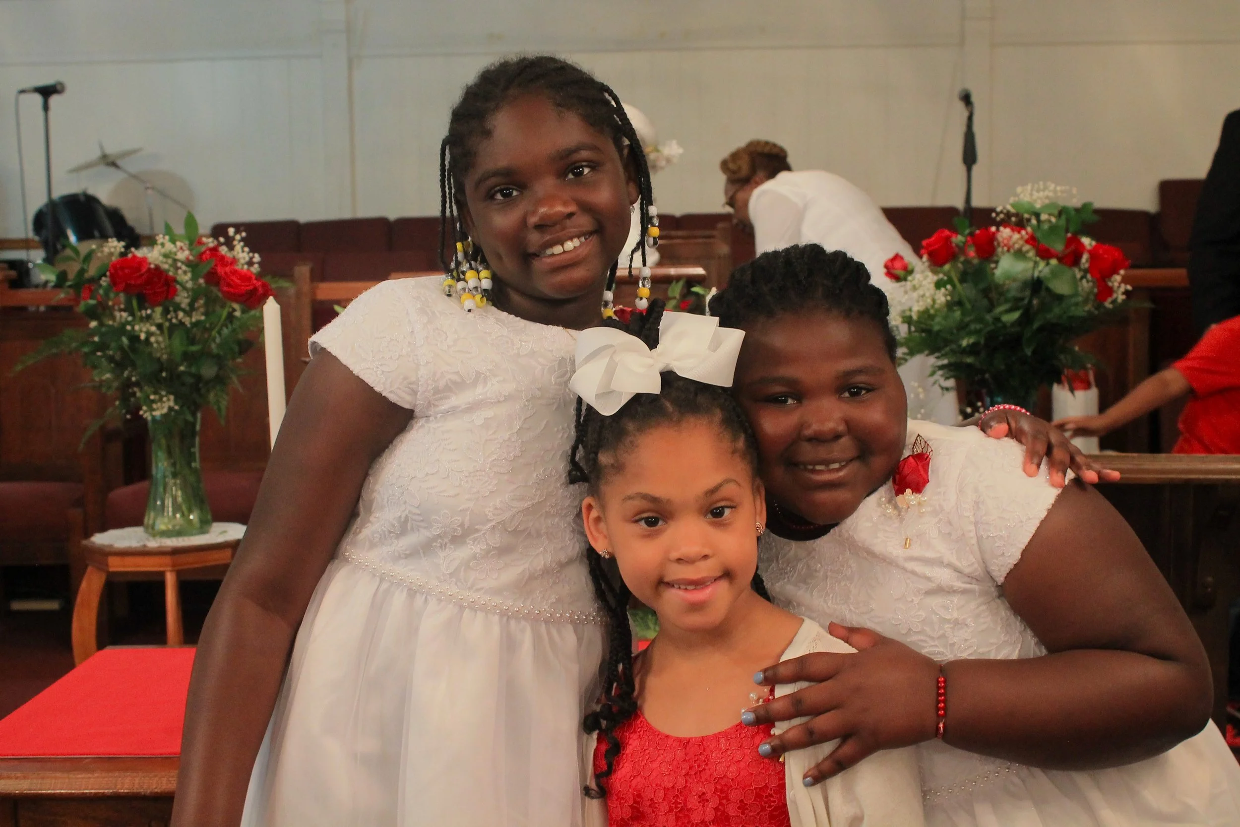 Three young girls dressed in white and red standing close together inside a church, with two large flower arrangements with red roses and white flowers in the background.